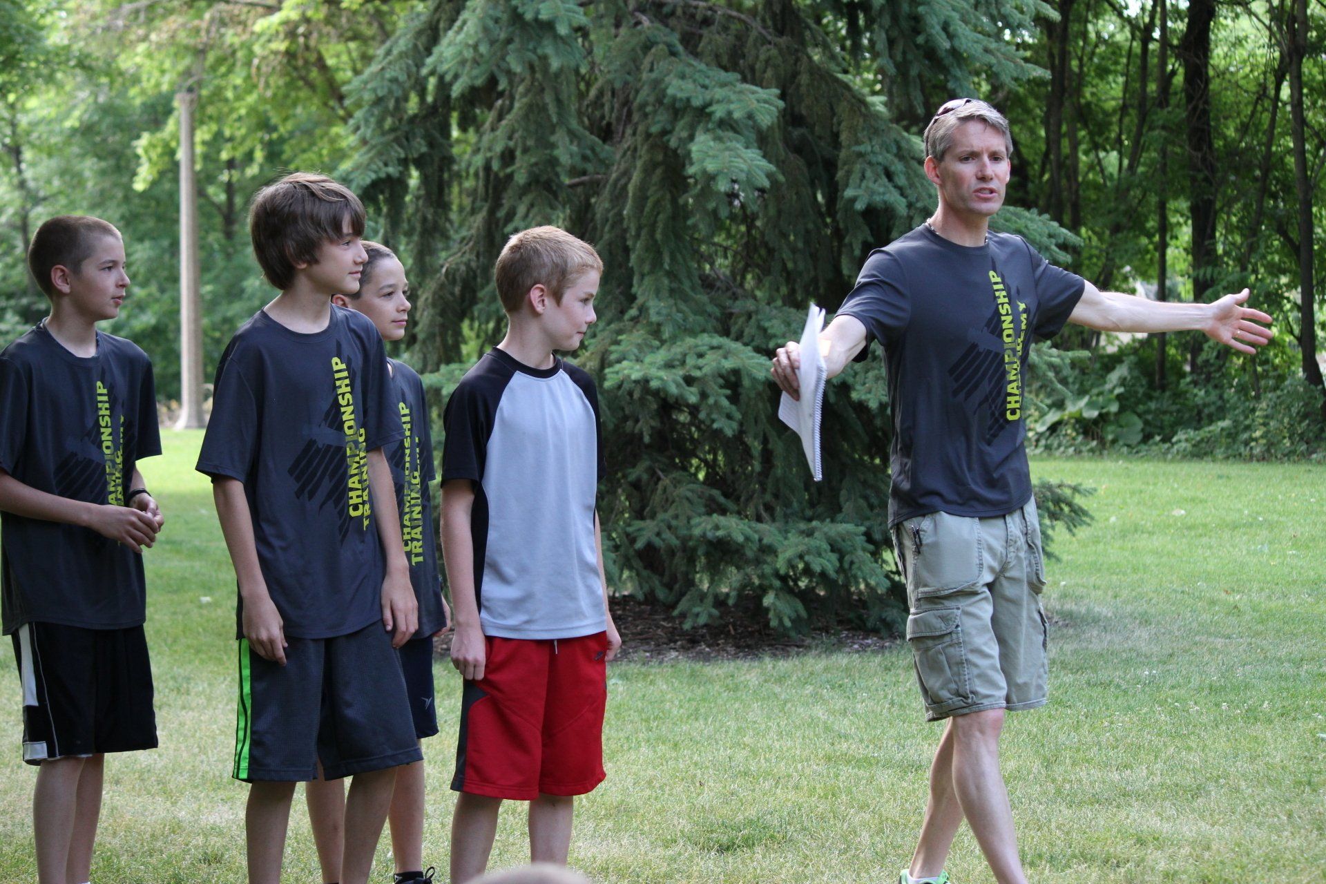 A group of young boys are standing in a grassy field while a man holds a piece of paper in his hand