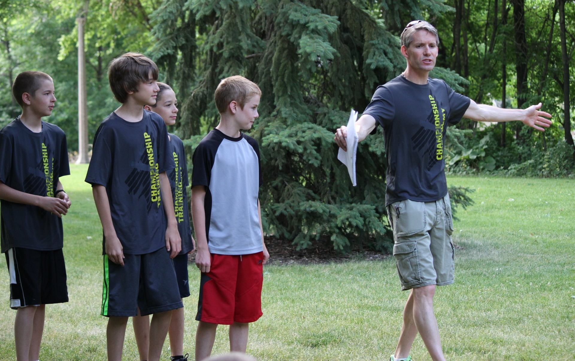 A group of young boys are standing in a grassy field while a man holds a piece of paper in his hand.
