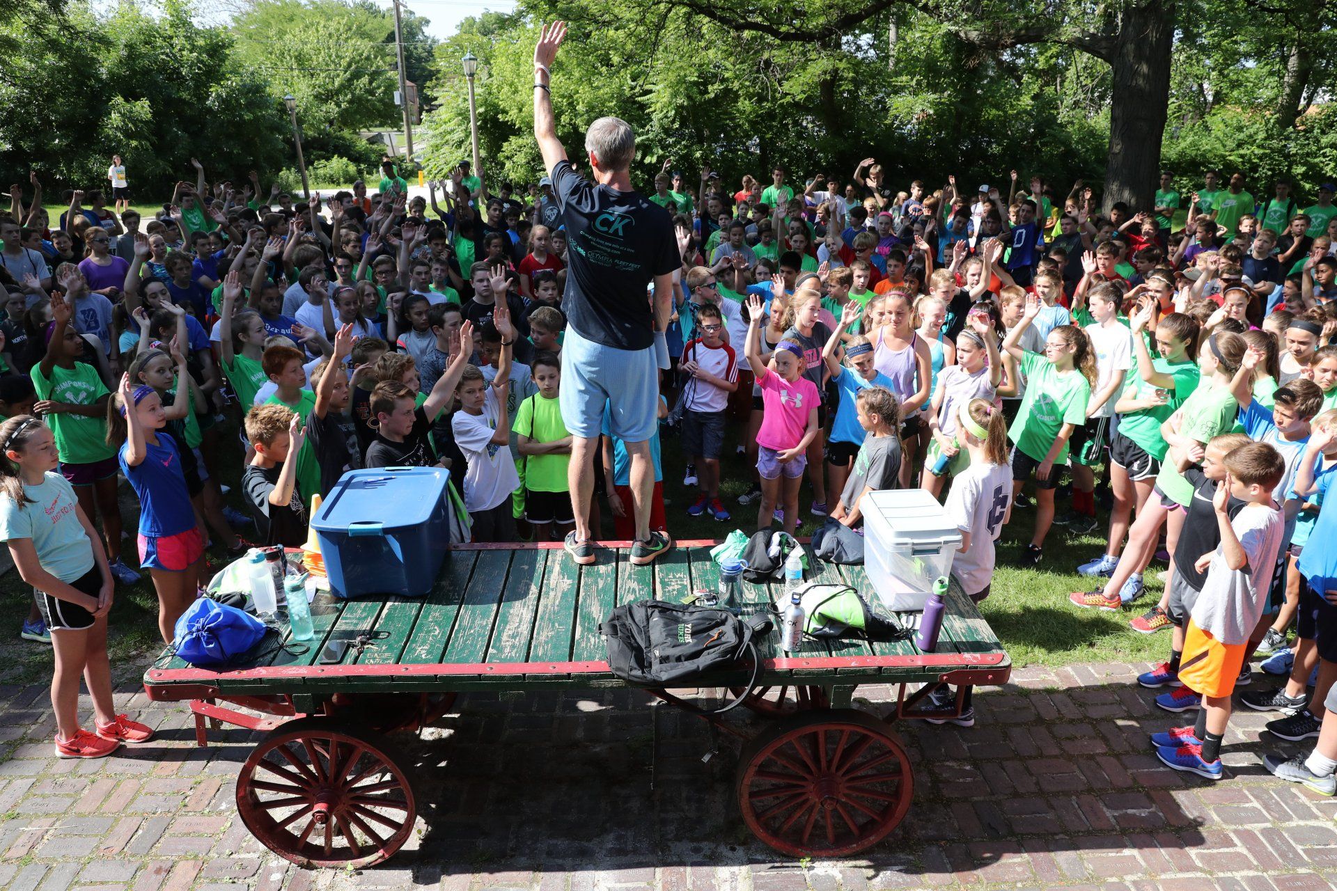 A man is standing on top of a wooden wagon in front of a crowd of children.