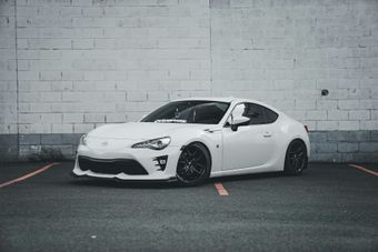 White sports car parked in front of a brick wall; black wheels and front lip.