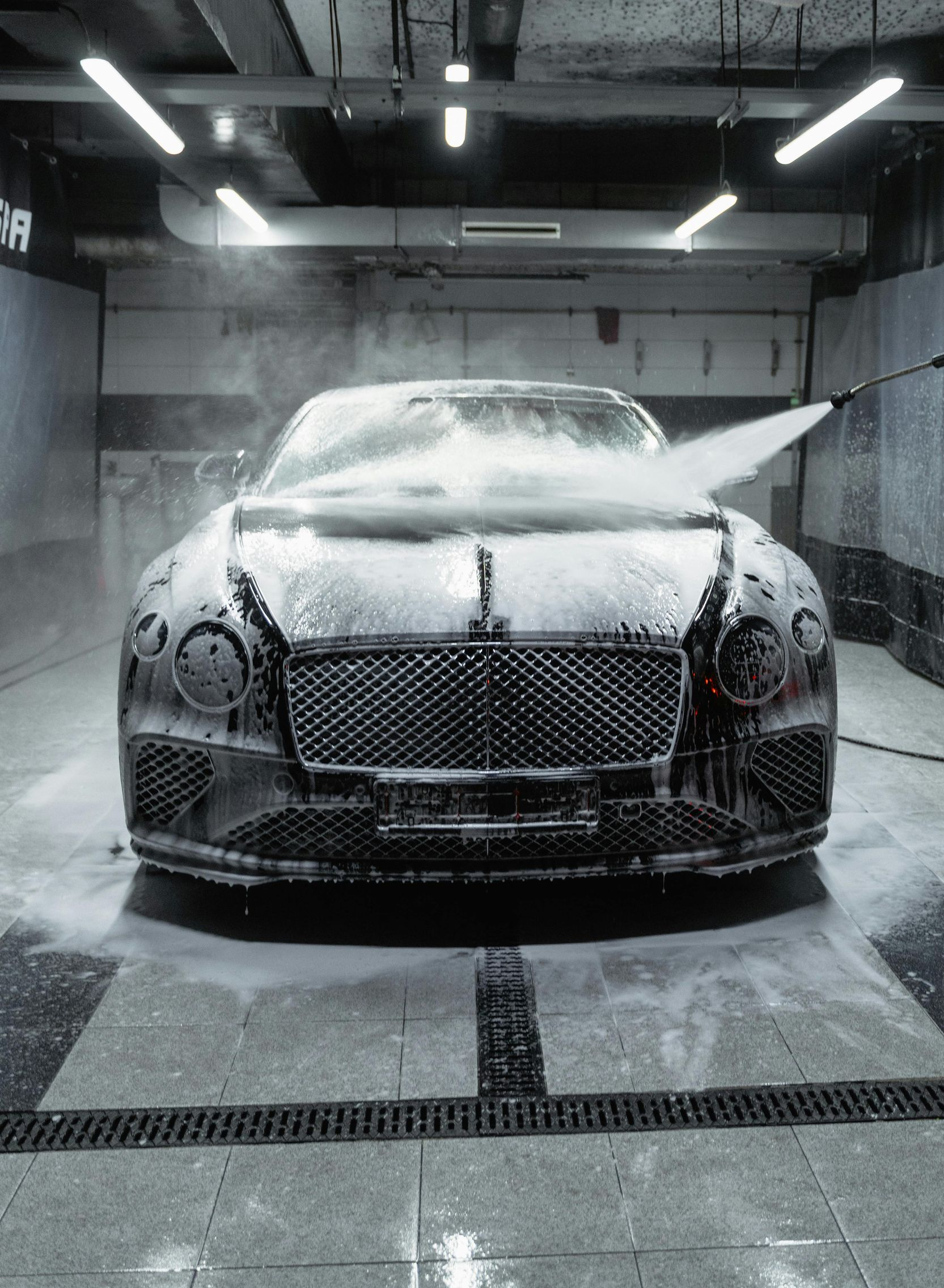 A Bentley car being washed at a car wash, covered in foam, spraying with high-pressure water.
