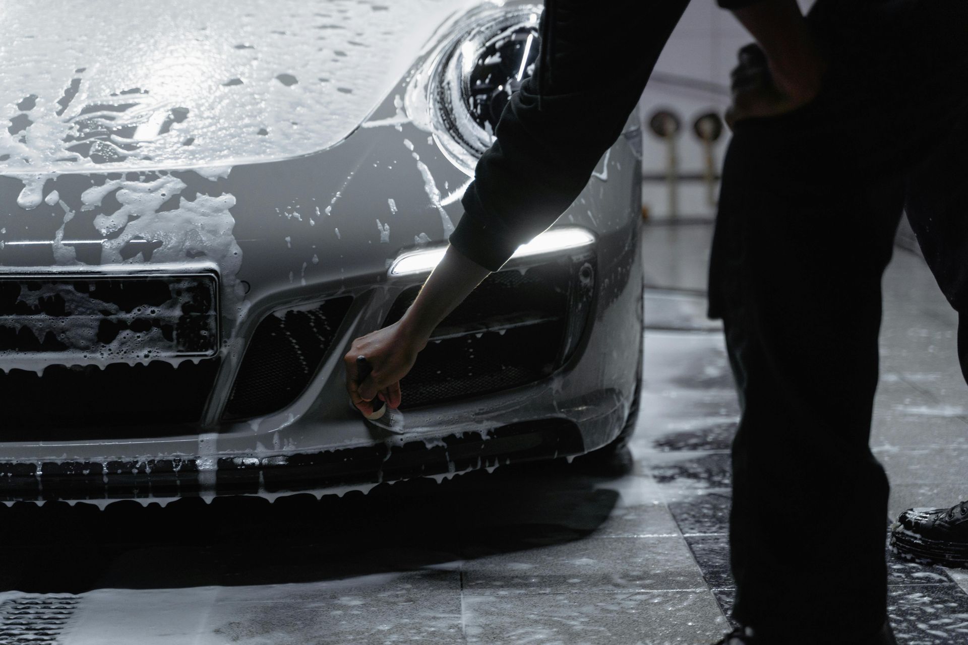 Person cleaning a gray sports car's bumper with a brush, covered in soap suds.
