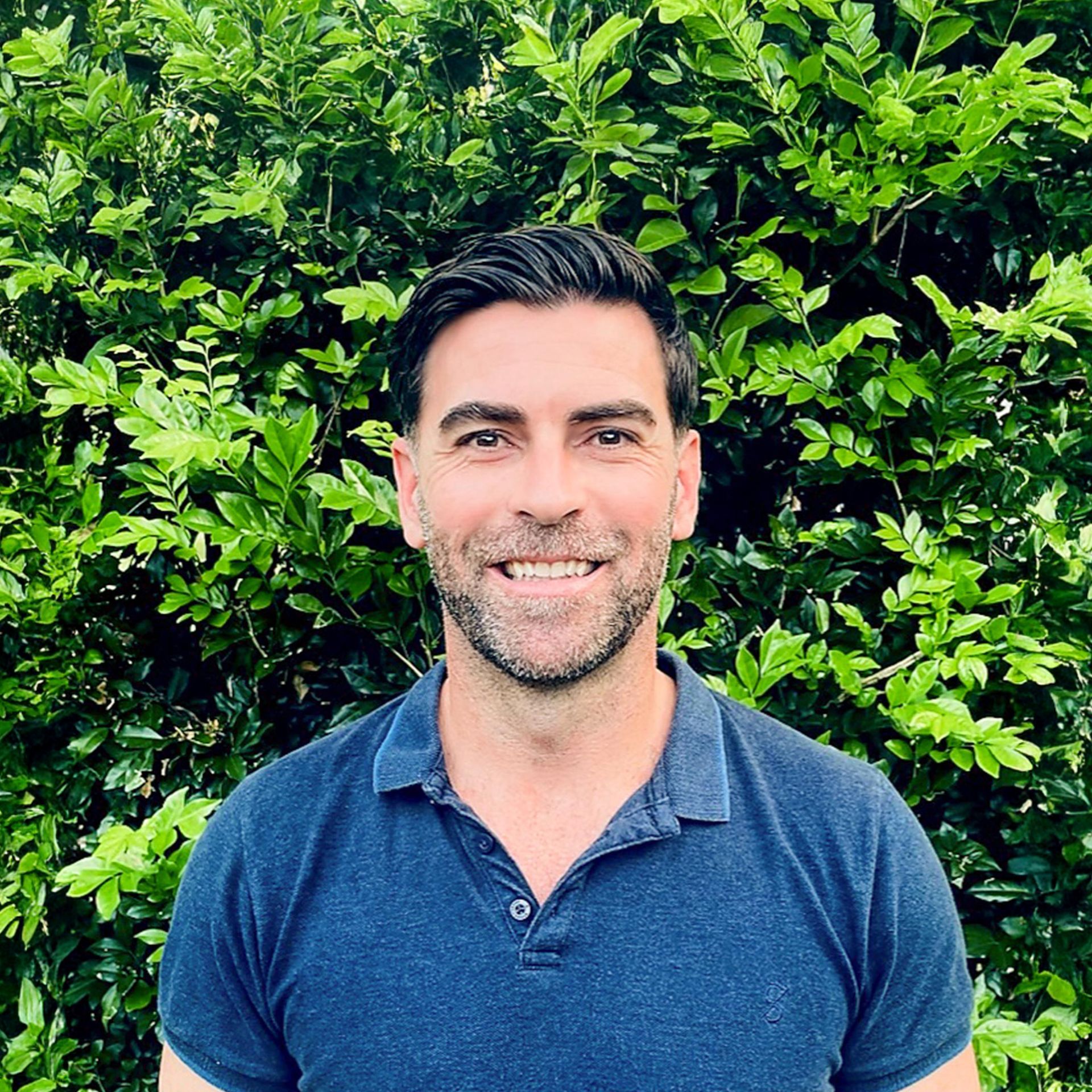 Izaac McClymont - Osteopath Bust shot of smiling man standing in front of shrubbery.