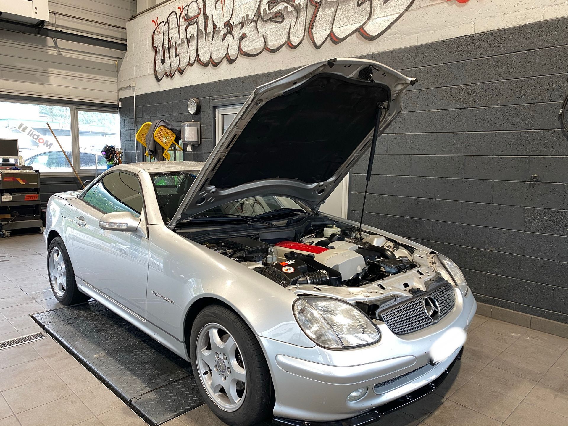 Une voiture argentée avec le capot relevé est garée dans un garage.