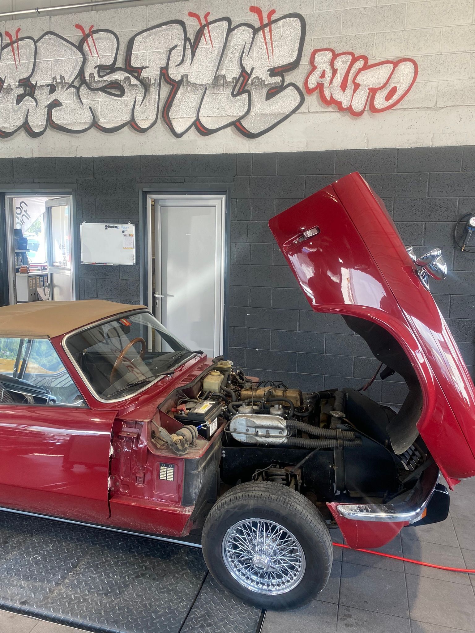 Une voiture rouge avec le capot relevé est garée devant un immeuble avec des graffitis dessus.