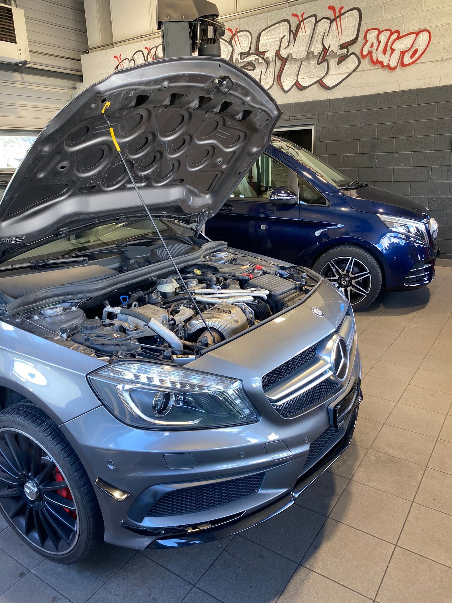 Une voiture avec le capot relevé est garée dans un garage à côté d’une autre voiture.