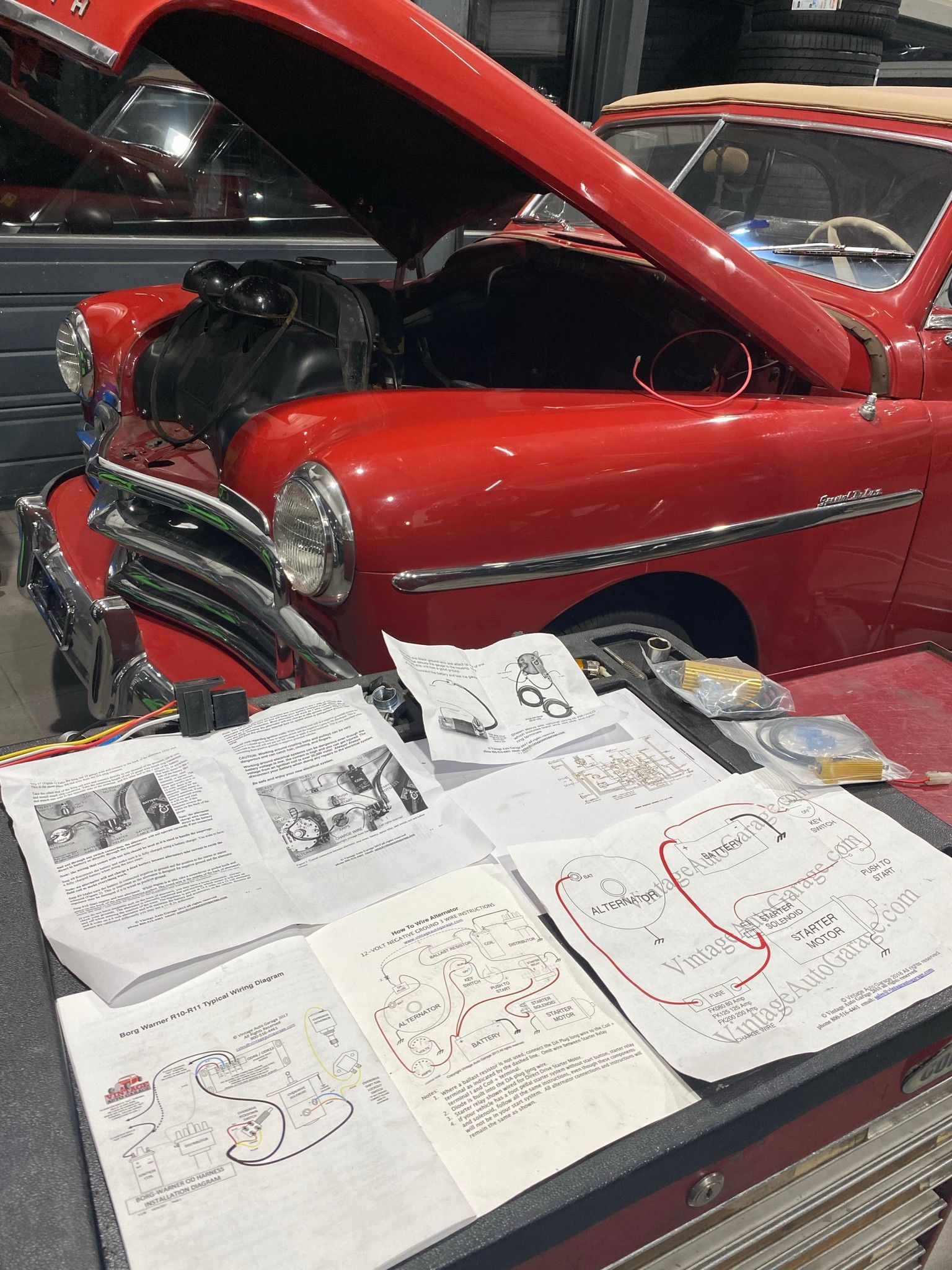 Une voiture rouge avec le capot relevé est garée à côté d’une table avec des papiers dessus.