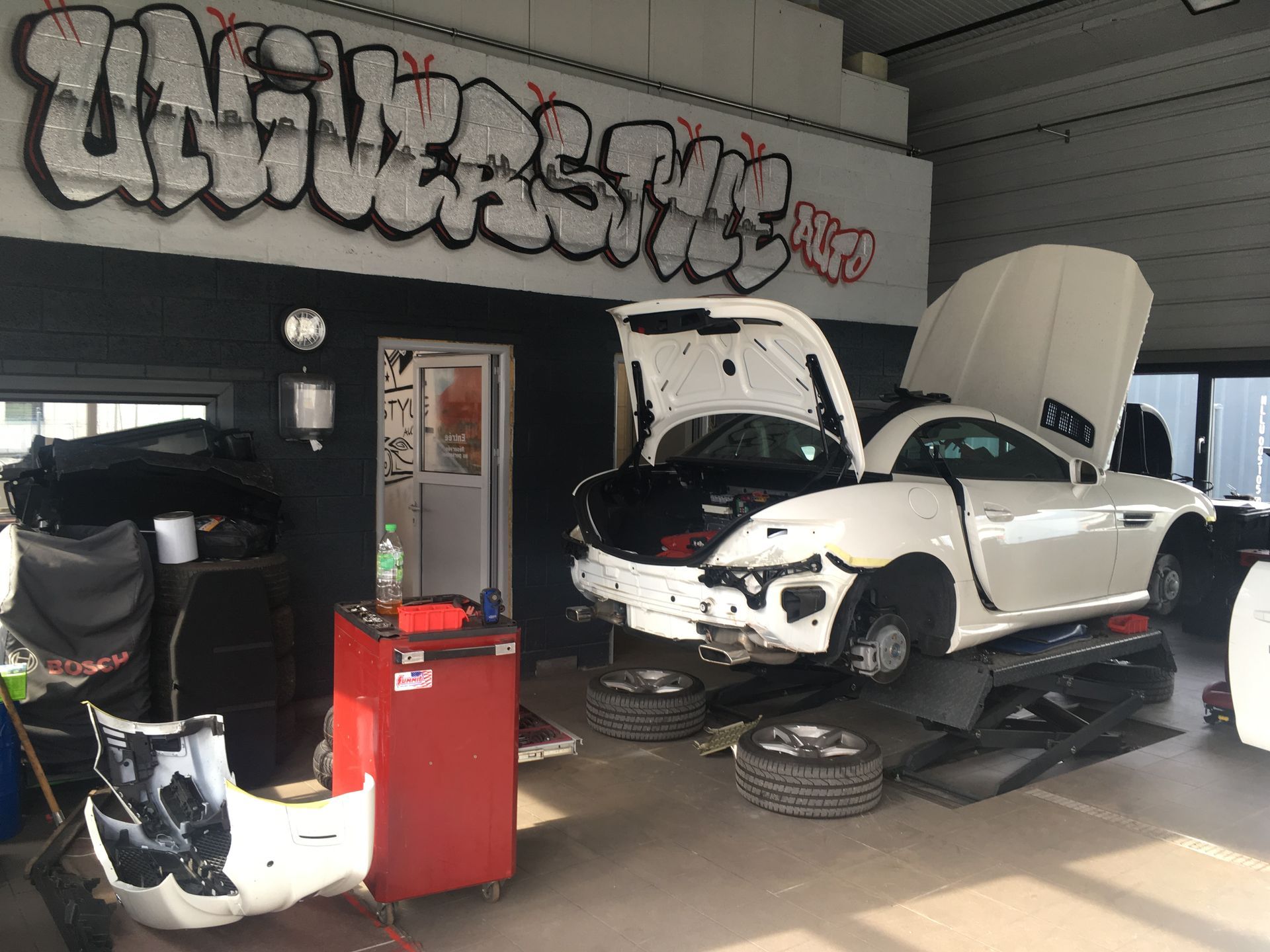 Une voiture blanche avec le capot ouvert se trouve dans un garage avec des graffitis sur le mur.