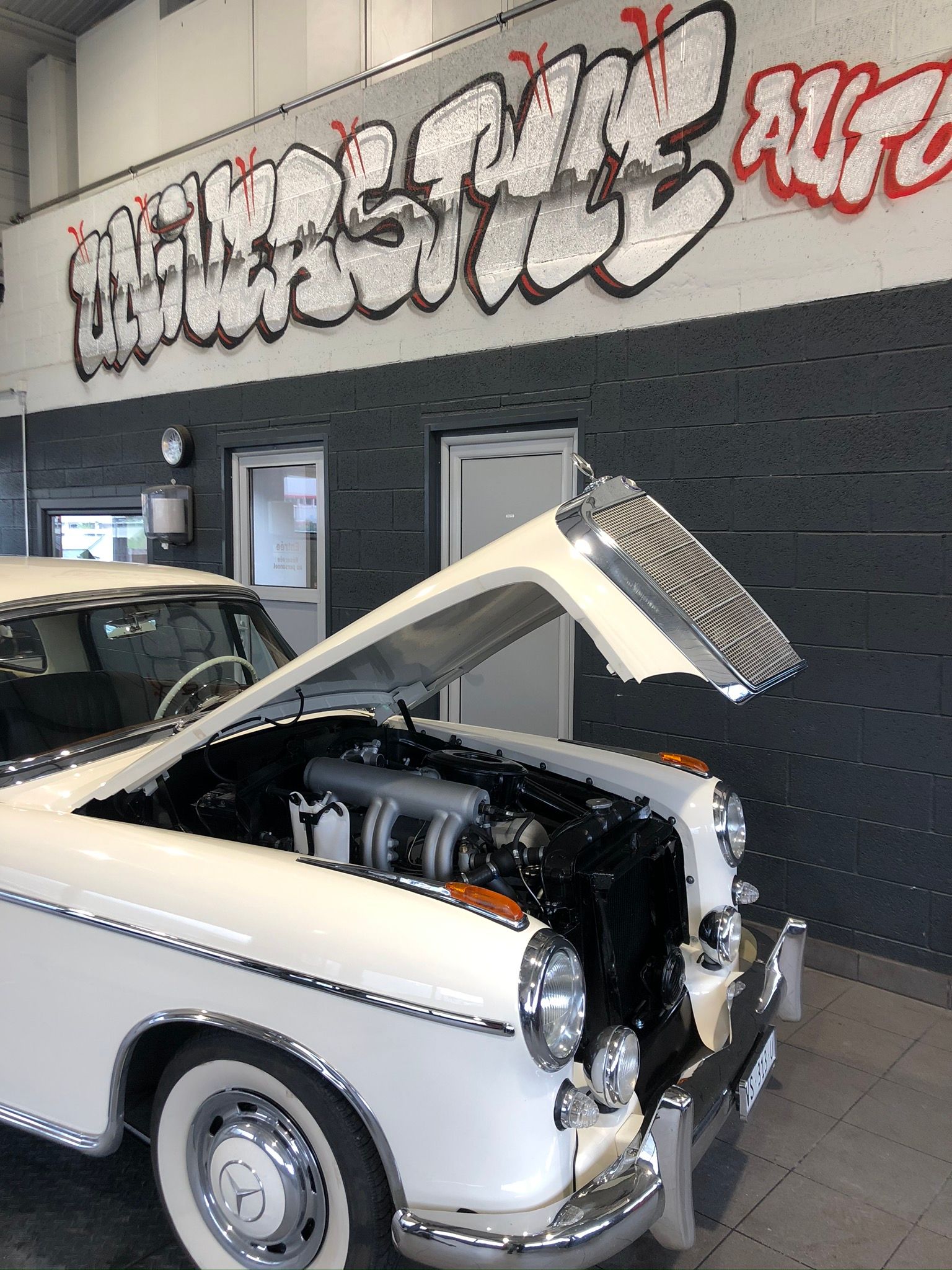 Une voiture blanche avec le capot ouvert est garée devant un mur couvert de graffitis.