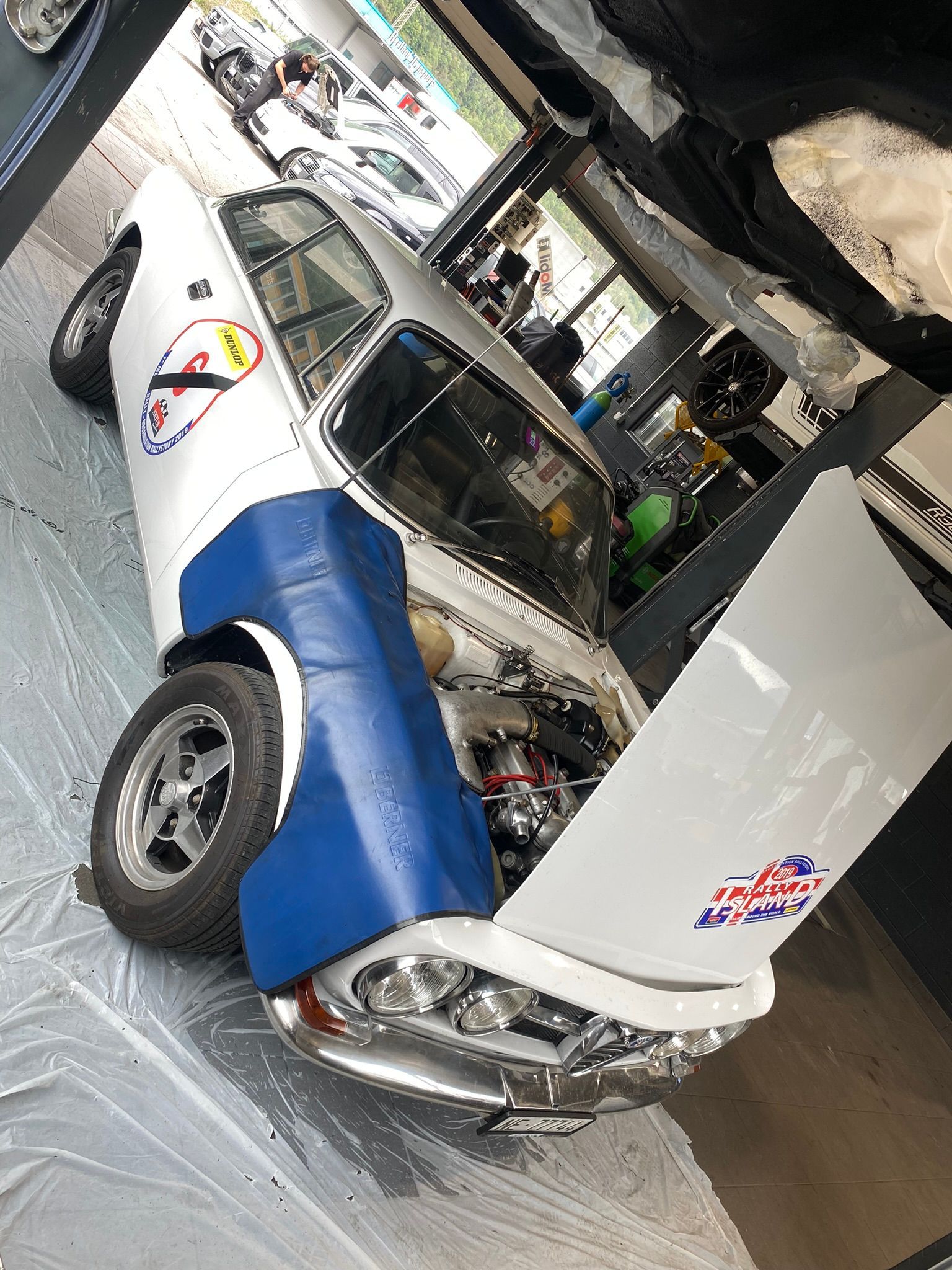 Une voiture blanche et bleue avec le capot ouvert dans un garage.