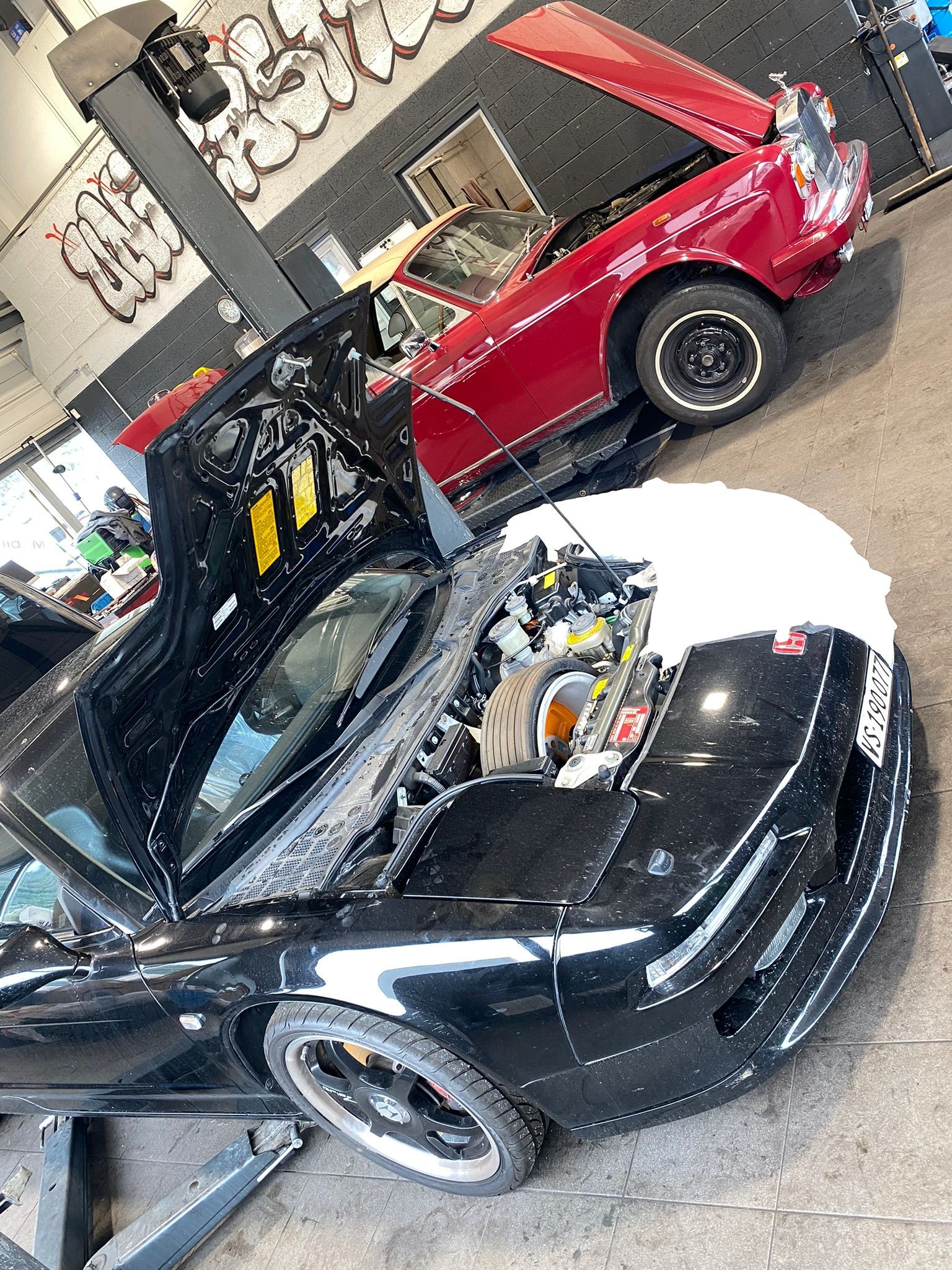 Une voiture noire avec le capot ouvert est garée à côté d’une voiture rouge dans un garage.