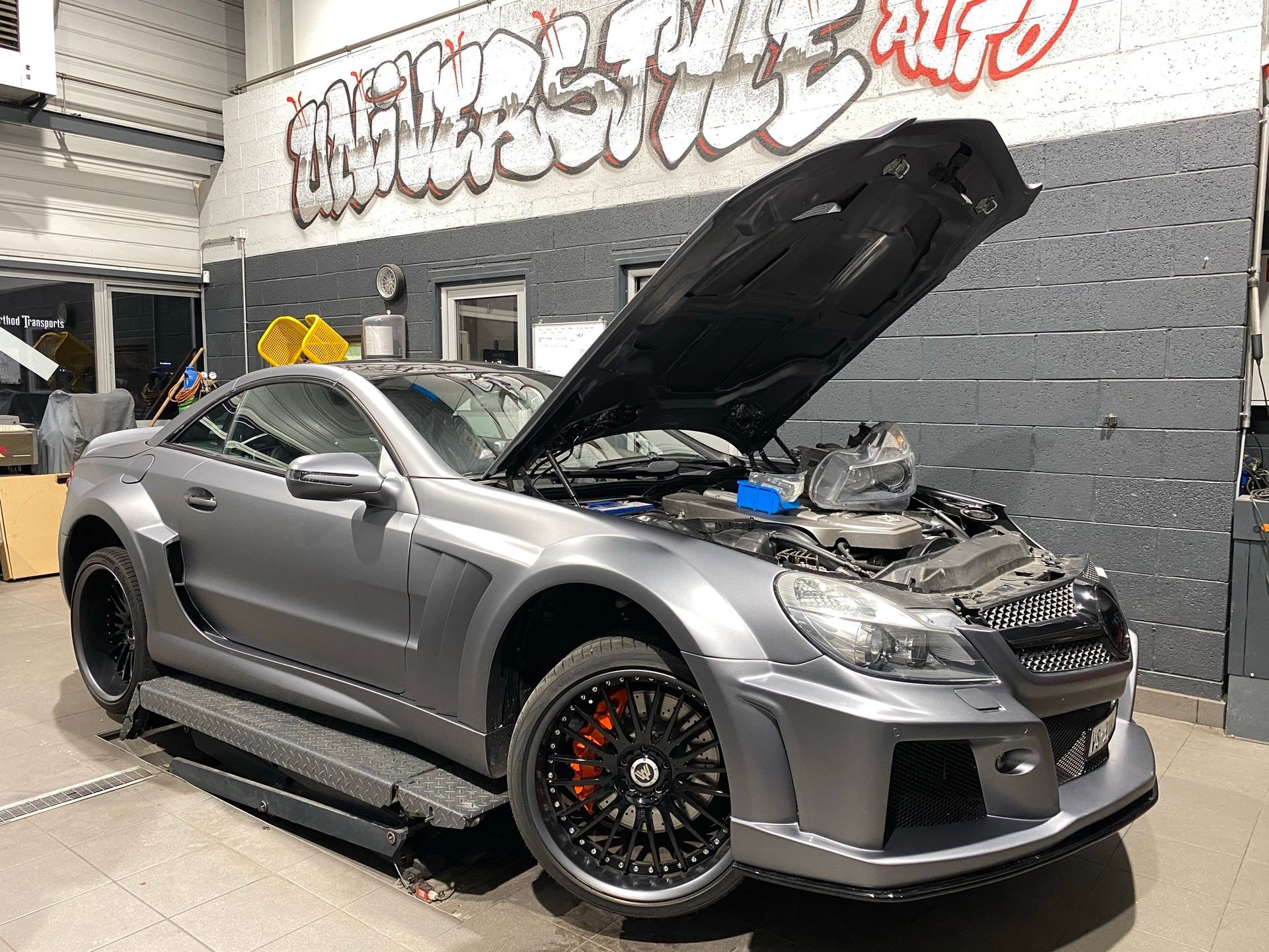 Une voiture de sport grise avec le capot relevé est garée devant un immeuble.