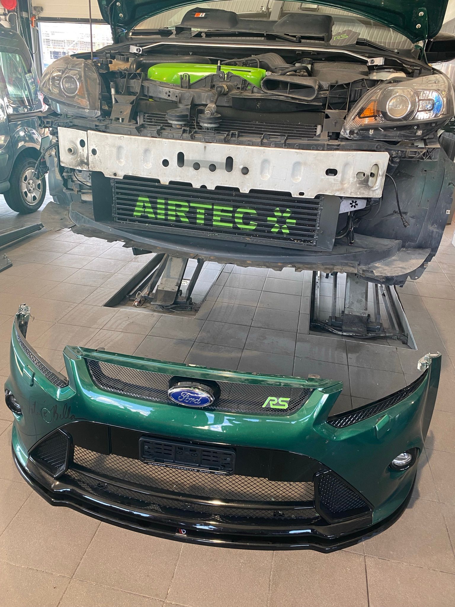 Une voiture verte dont le pare-chocs avant a été retiré est assise sur un ascenseur dans un garage.