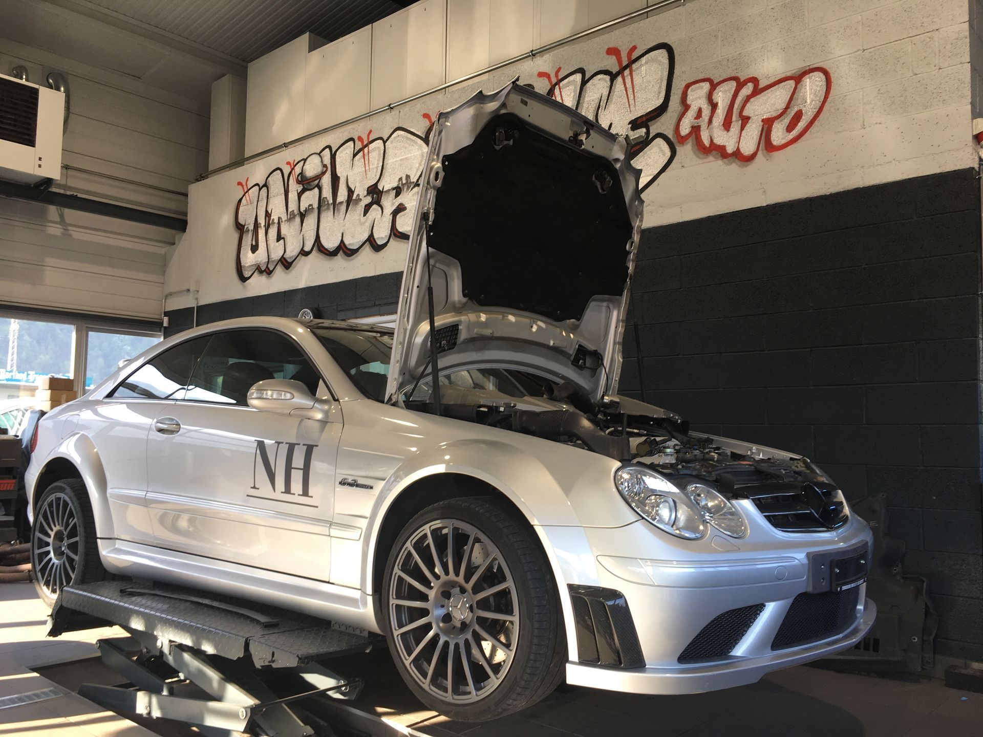 Une voiture argentée avec le capot ouvert est posée sur un ascenseur dans un garage.