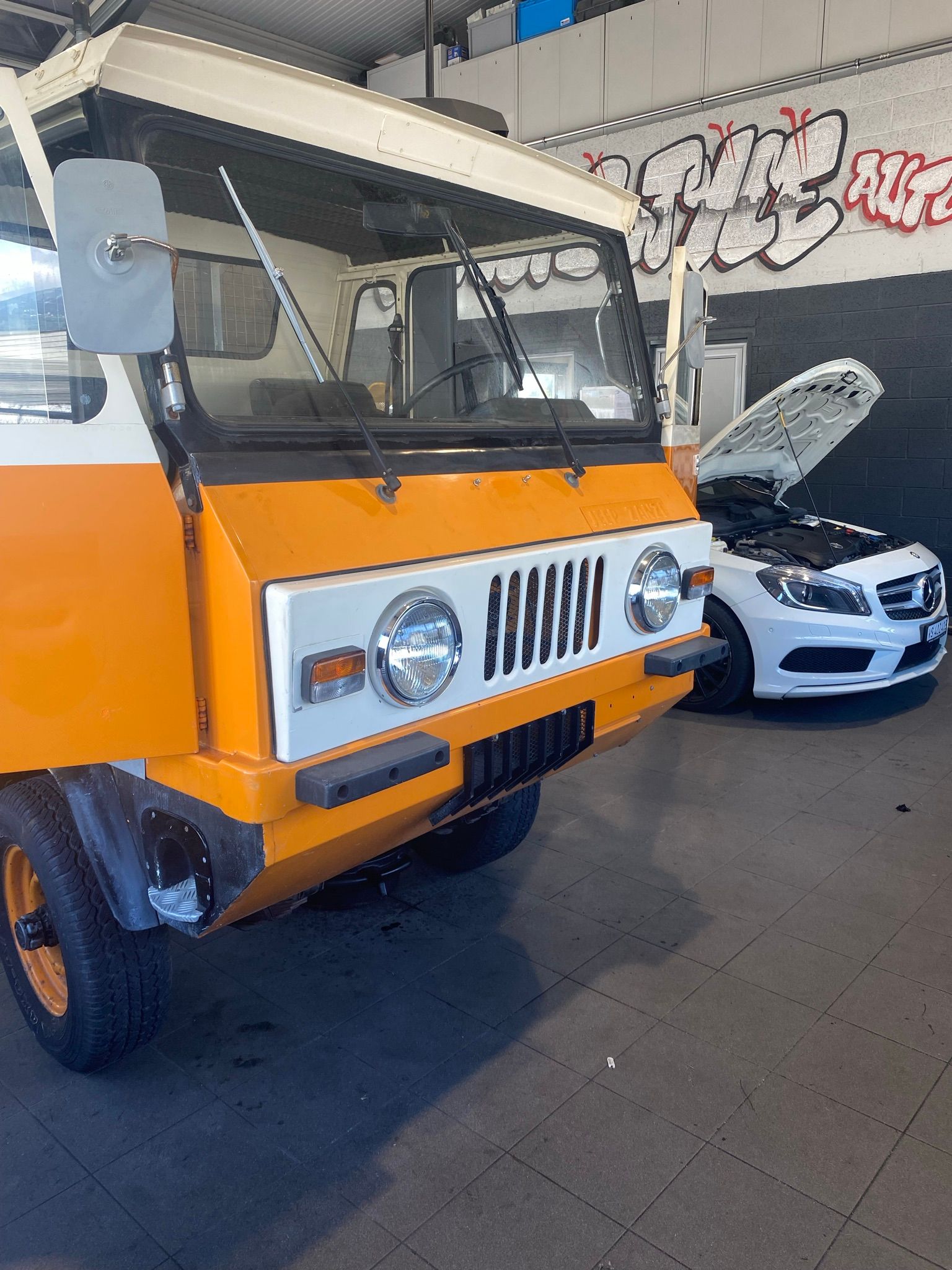 Un véhicule orange et blanc est garé à côté d’une voiture blanche avec le capot ouvert.