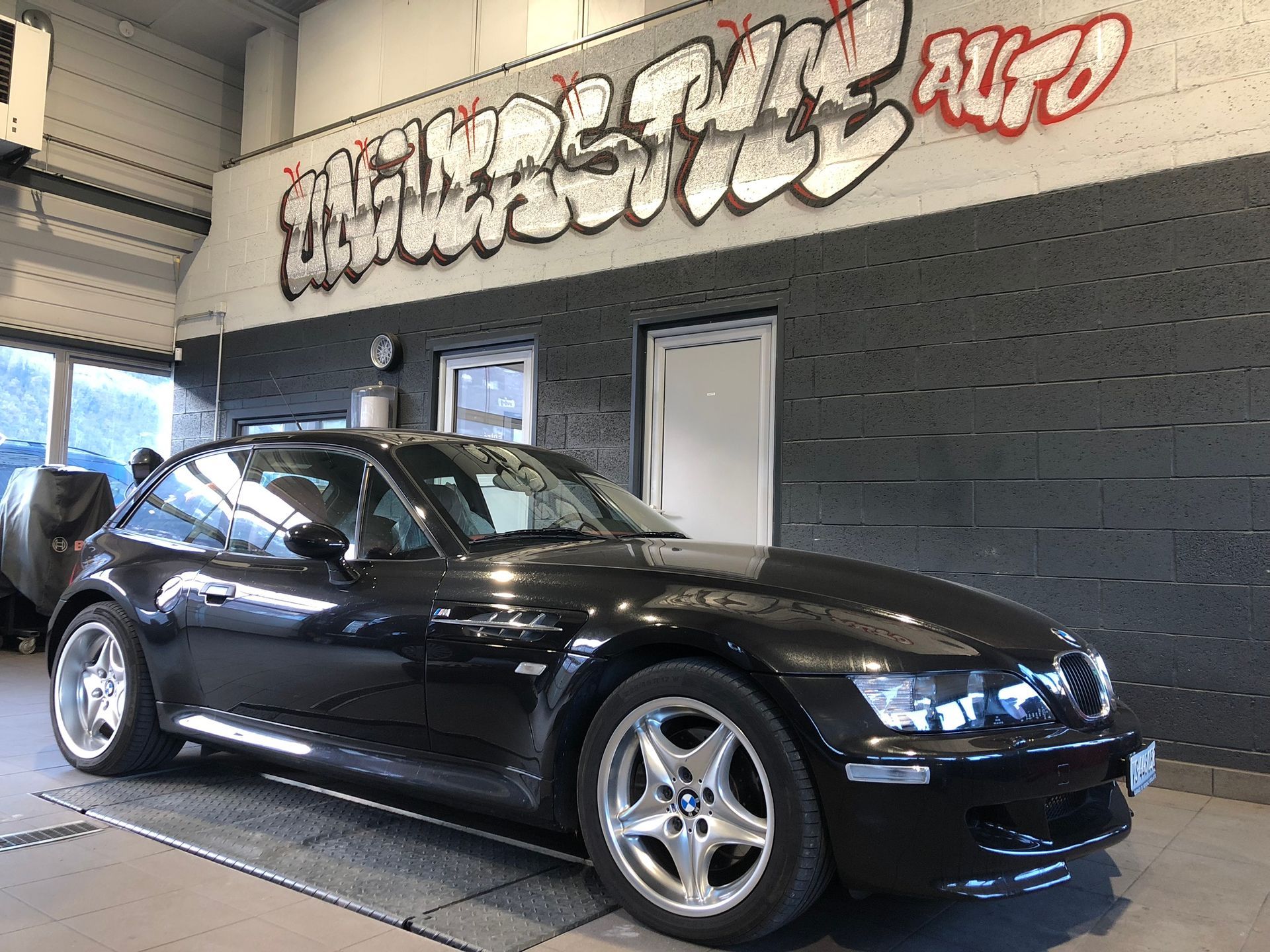 Une BMW Z3 noire est garée devant un mur couvert de graffitis.