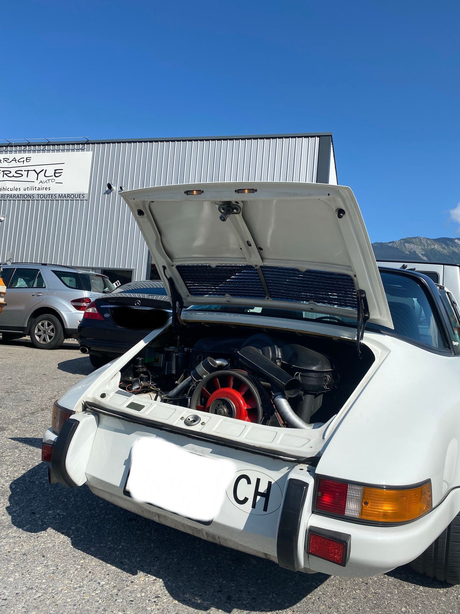 Une Porsche blanche avec le capot relevé est garée dans un parking.