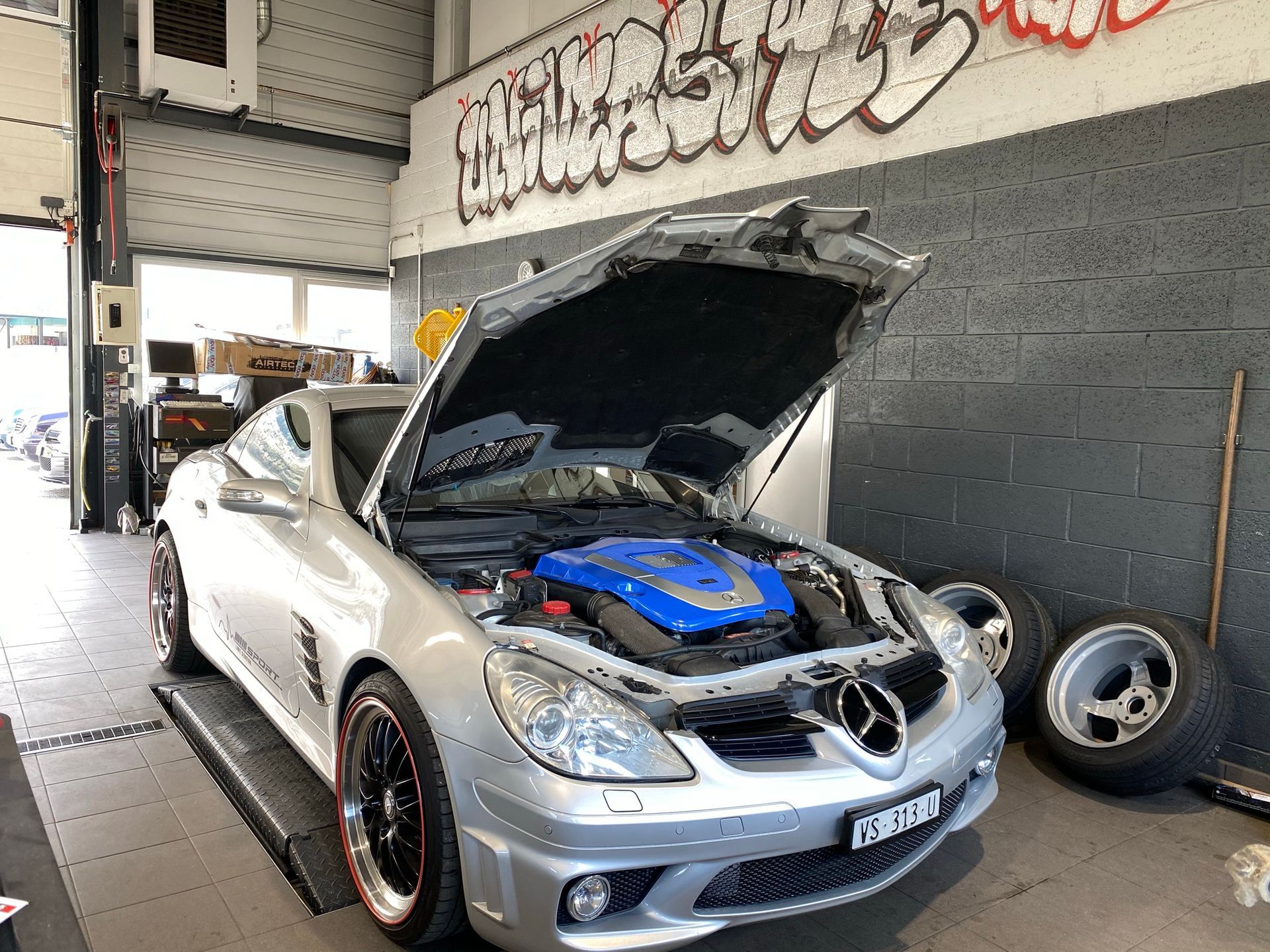 Une voiture argentée avec le capot relevé se trouve dans un garage.