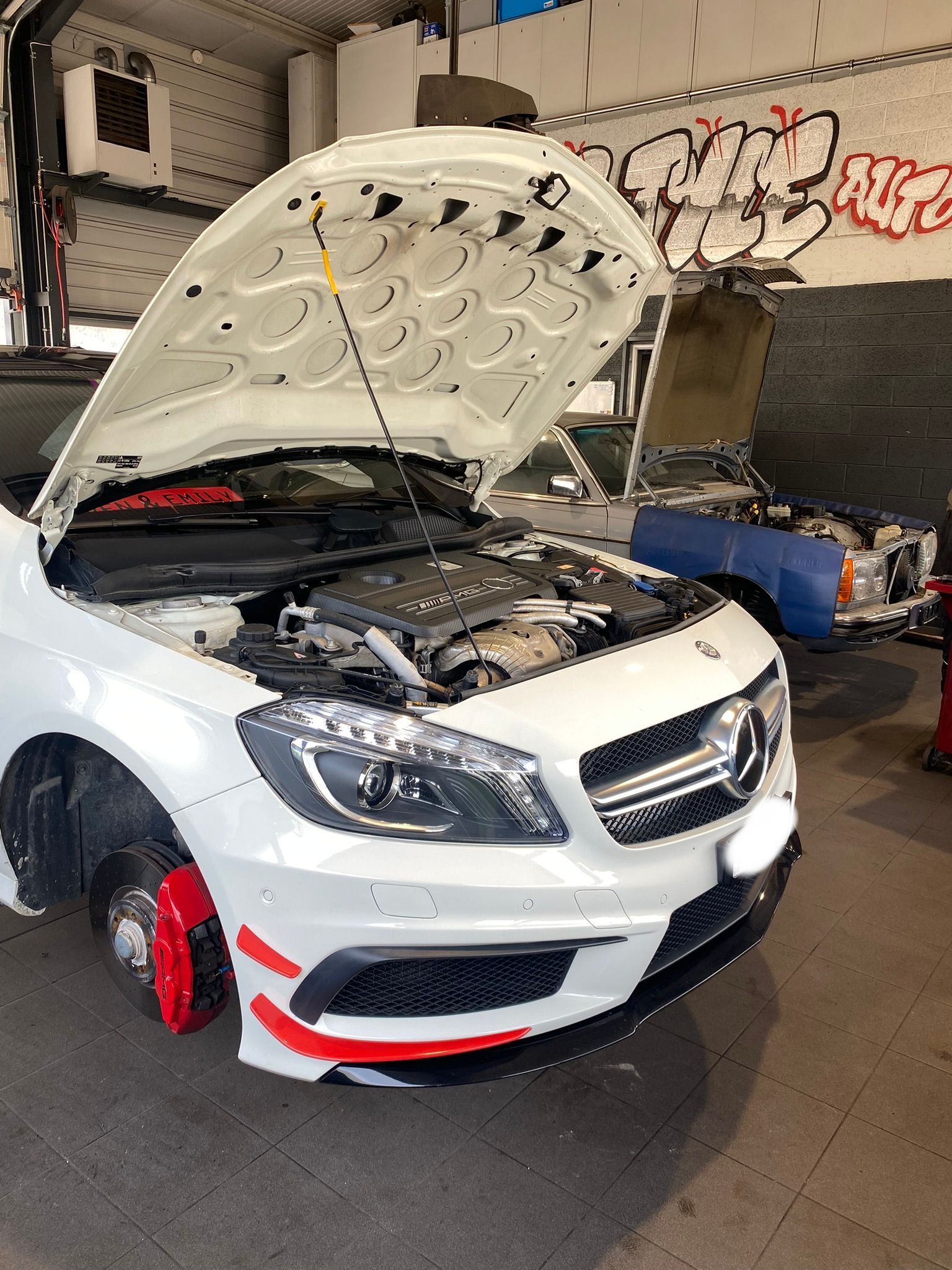 Une Mercedes Benz blanche avec le capot ouvert dans un garage.