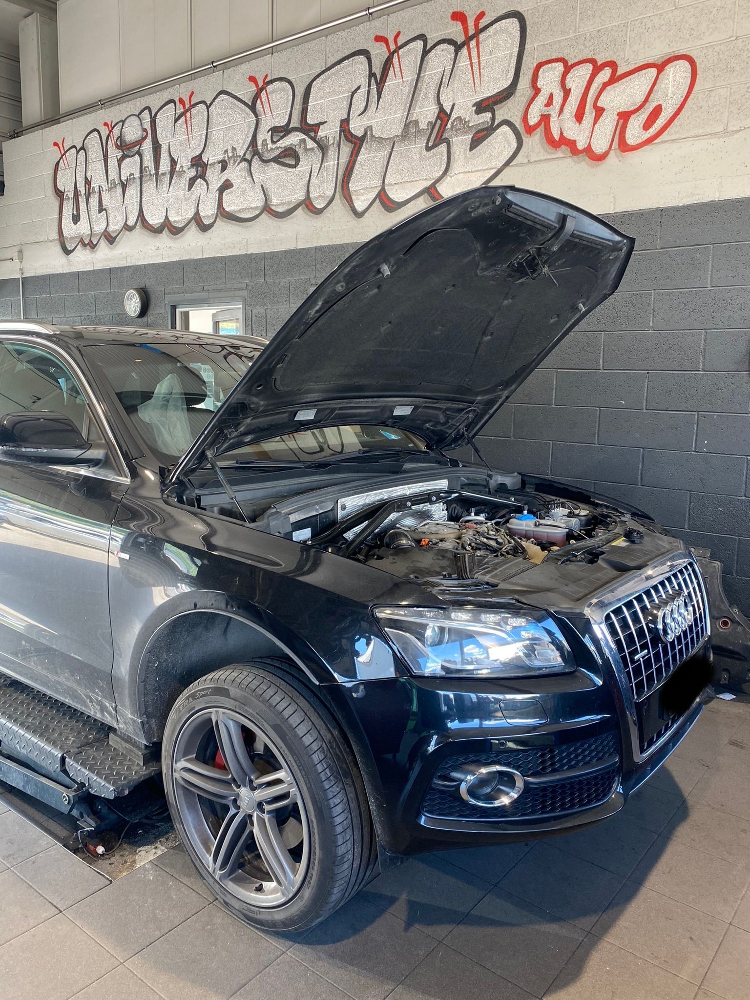 Une voiture noire avec le capot relevé est garée devant un mur avec des graffitis dessus.