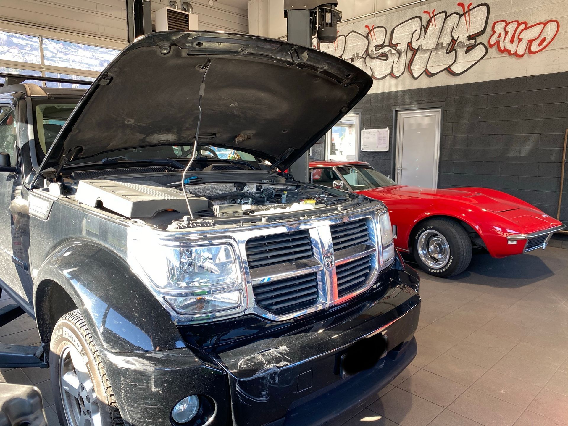 Une Dodge Nitro noire avec le capot ouvert est garée dans un garage à côté d'une corvette rouge.