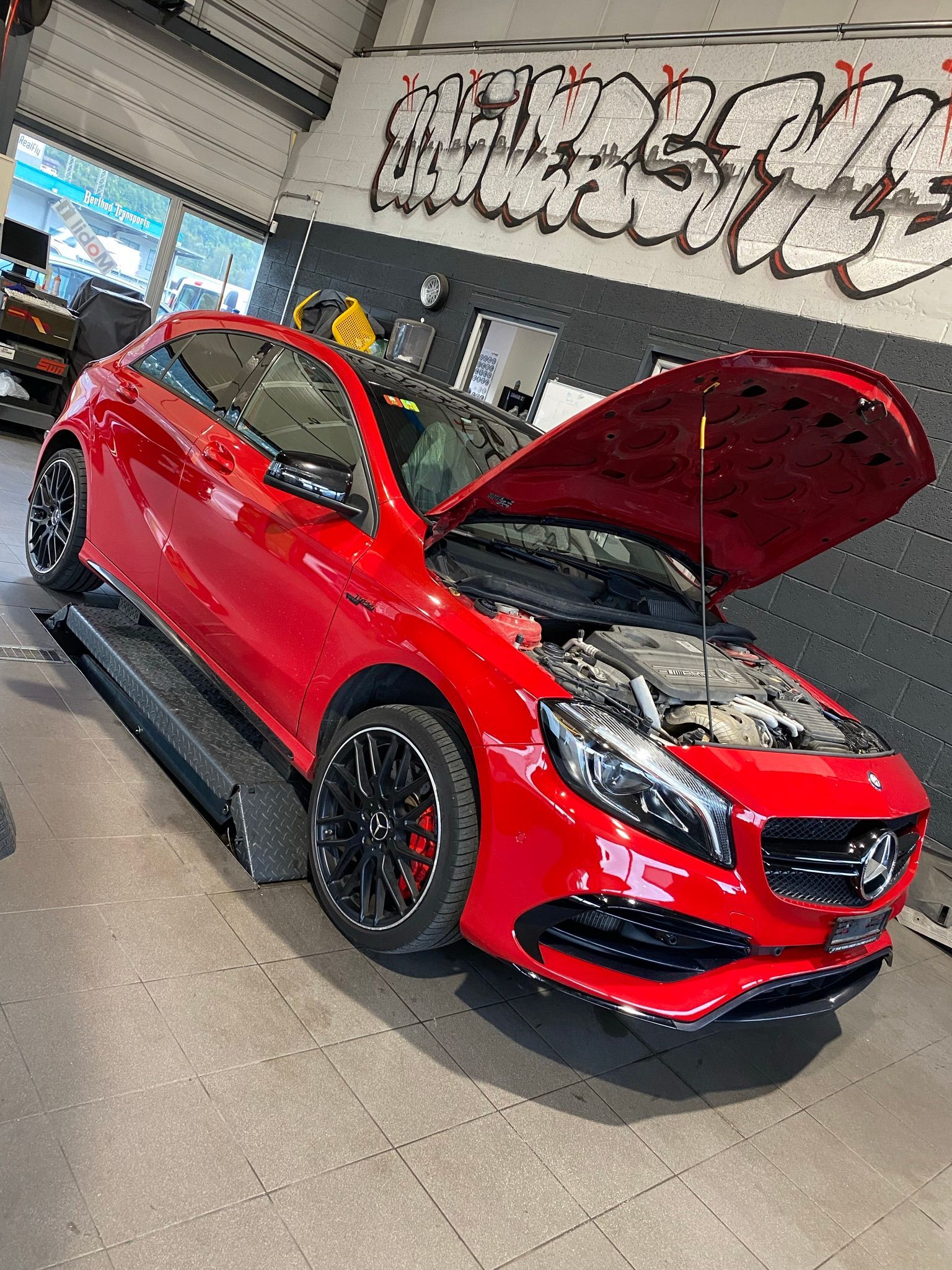 Une Mercedes Benz rouge avec le capot ouvert est garée dans un garage.