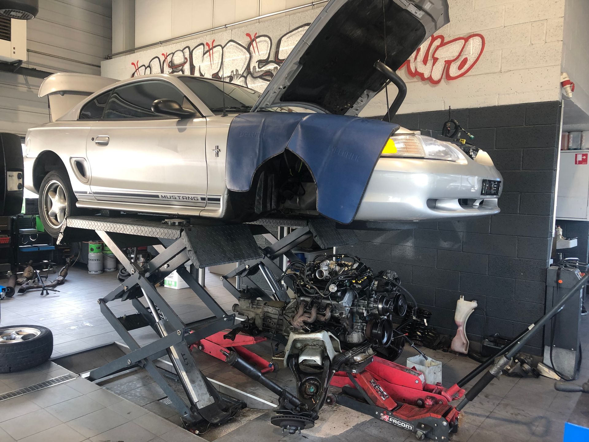 Une voiture argentée est assise sur un ascenseur dans un garage avec le capot relevé.