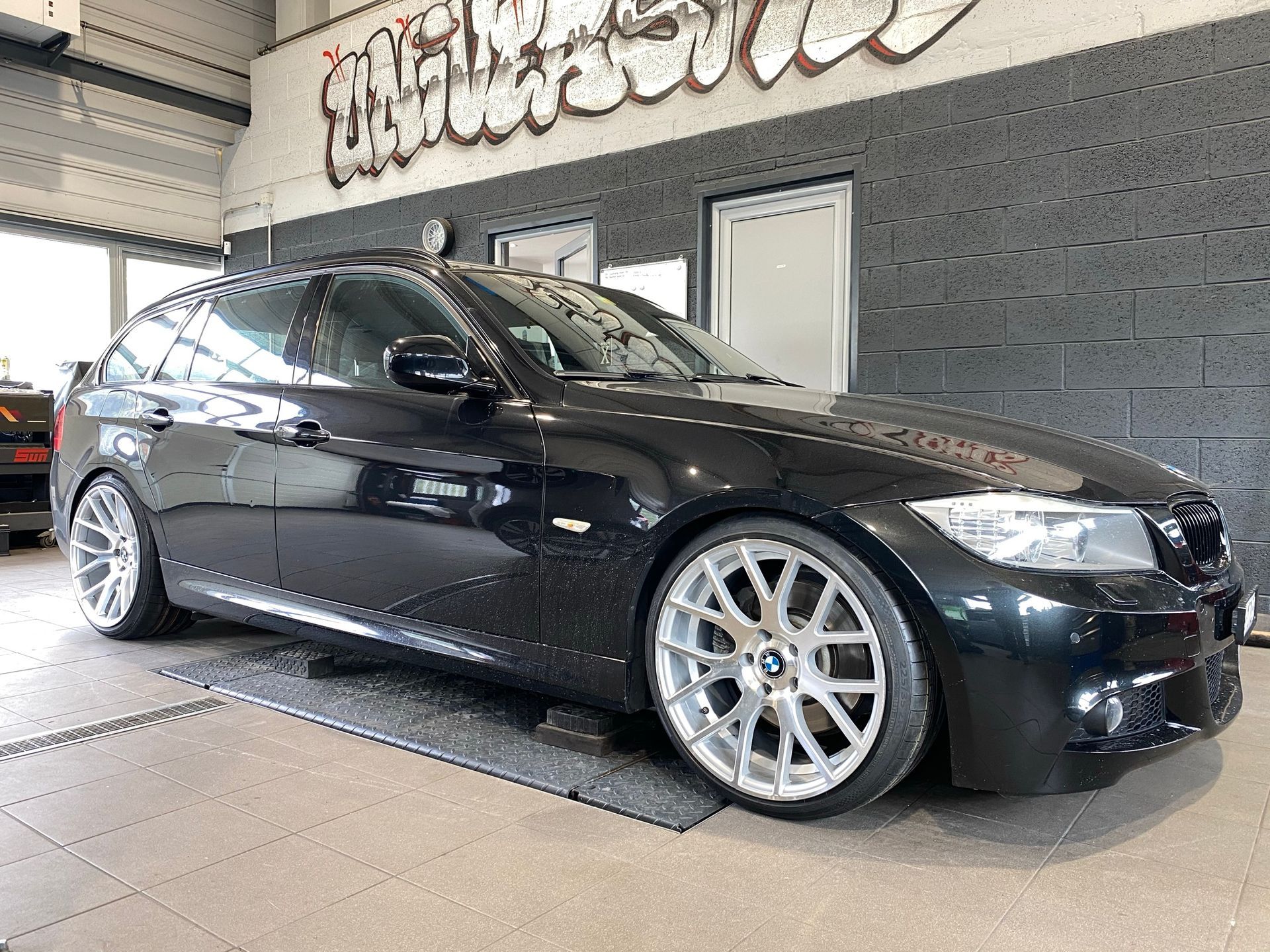 Une BMW Série 3 noire est garée dans un garage avec des graffitis sur le mur.