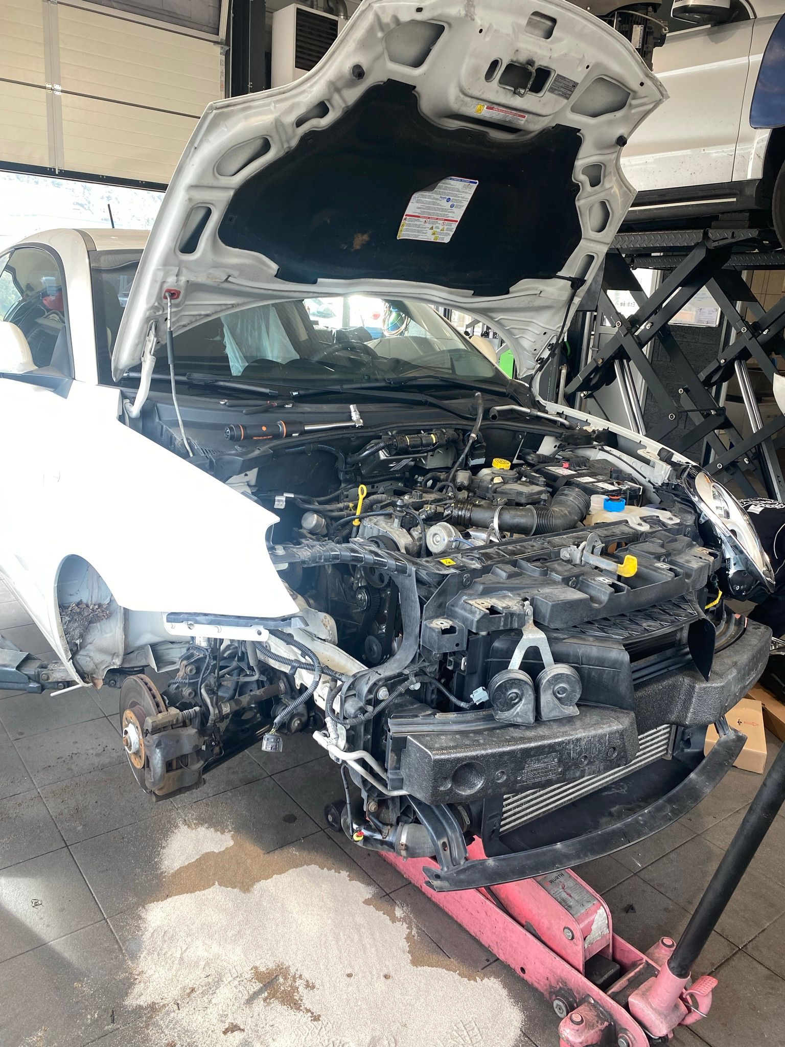 Une voiture blanche avec le capot ouvert est posée sur un cric rose dans un garage.