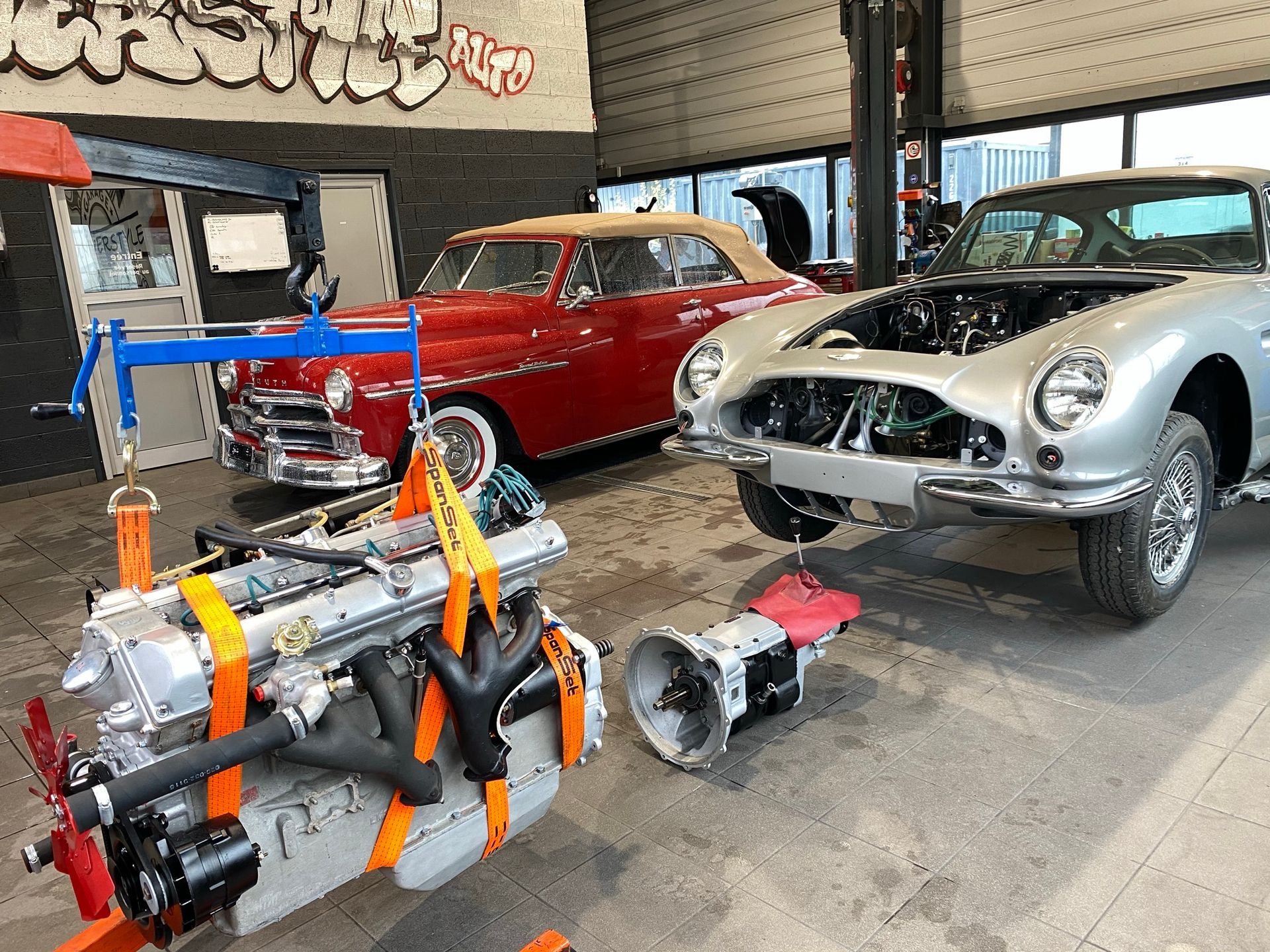 Une voiture argentée est en cours de réparation dans un garage à côté d'une voiture rouge.