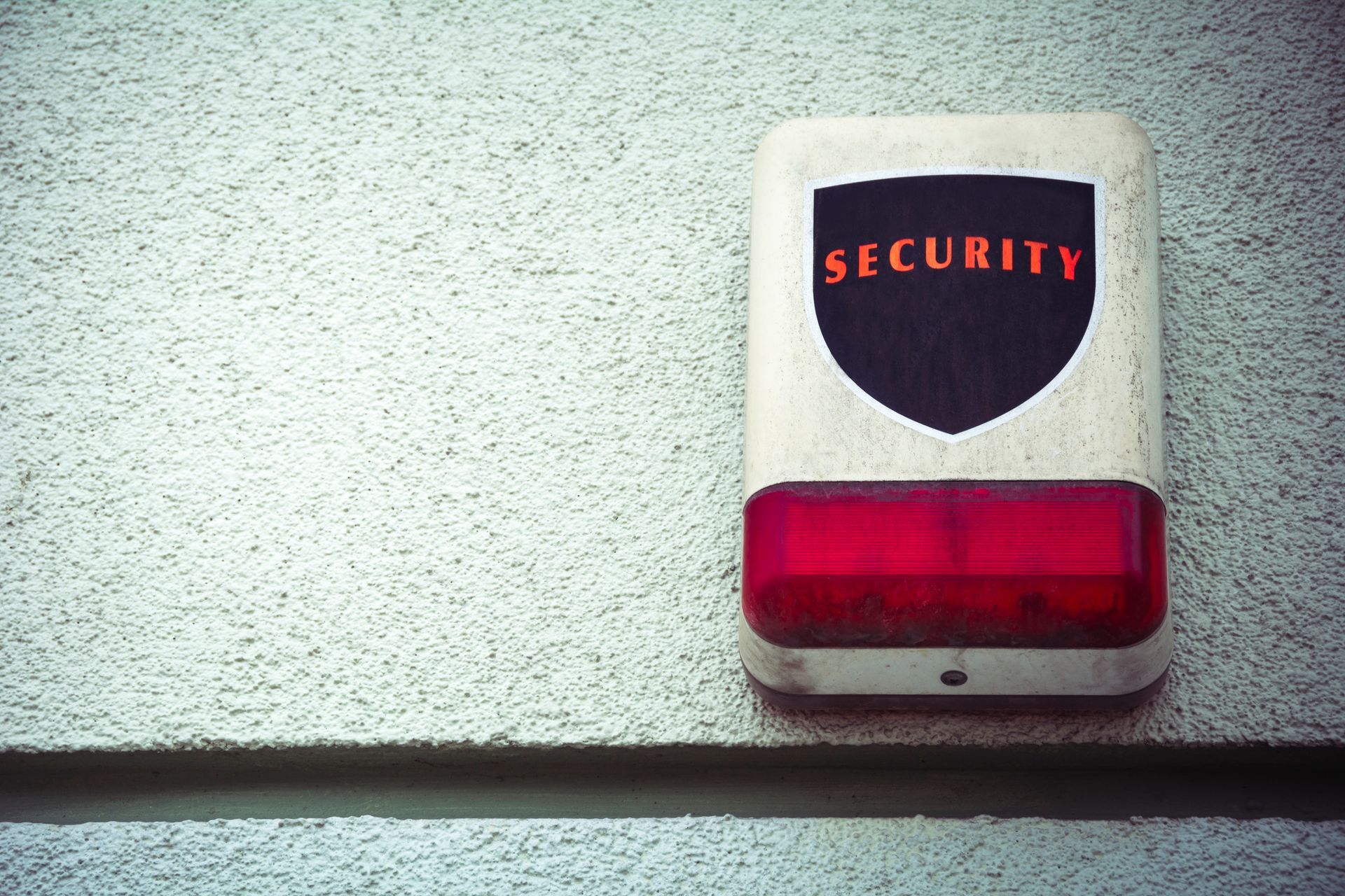 Security alarm on a textured white wall; red light and black/orange shield graphic with 