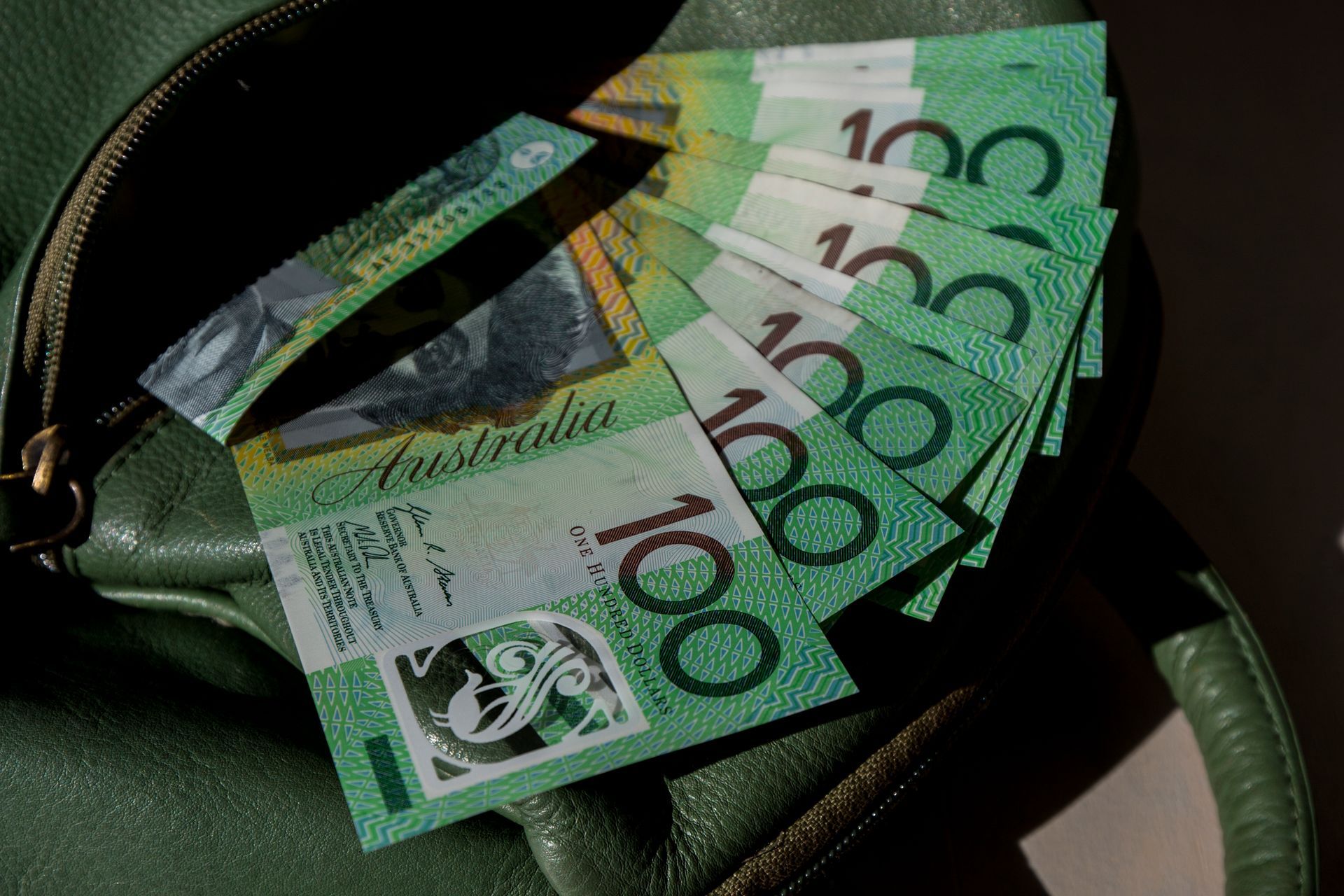 Green Australian $100 bills fanned out from a dark green bag.