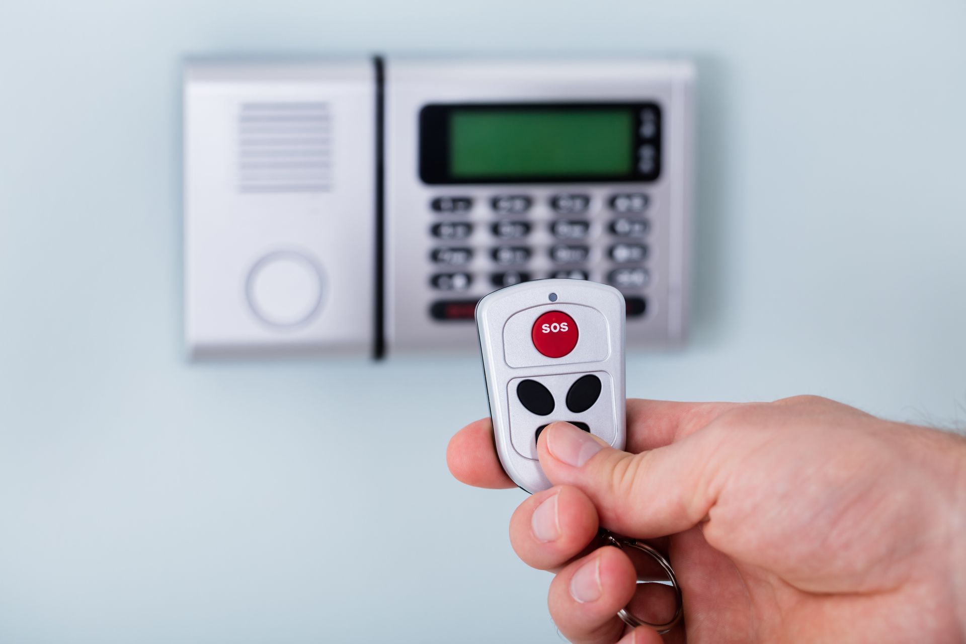 Hand holding a white remote, pointing it at a security system keypad on a light blue wall.