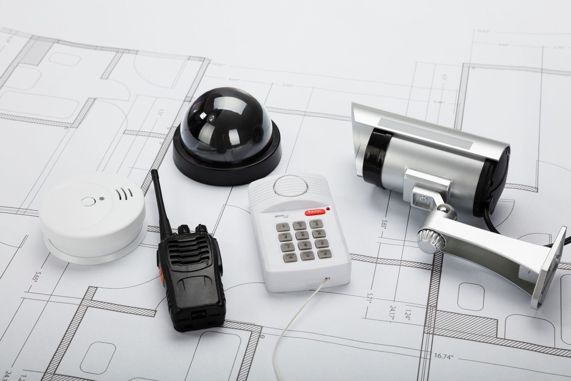 Security system components on a blueprint: dome camera, surveillance camera, keypad, walkie-talkie, and smoke detector.
