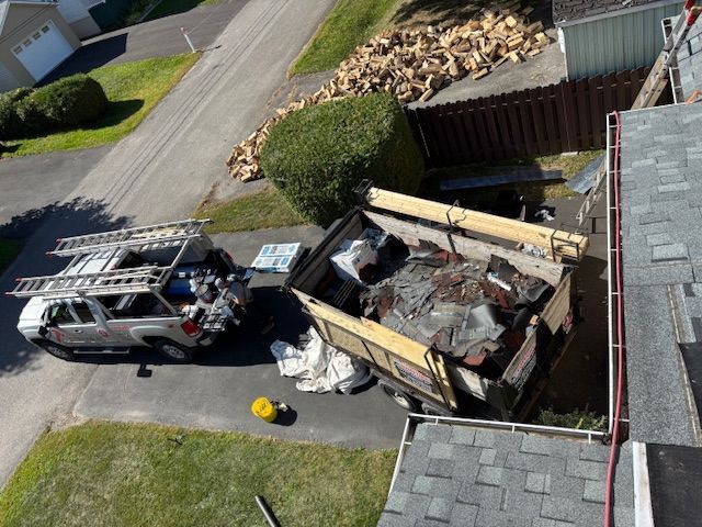 Vue aérienne : benne à ordures en cours de remplissage avec des matériaux de toiture ; SUV avec échelle sur le dessus, stationné à proximité dans une rue résidentielle.