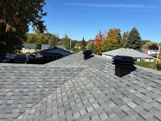 Toit en bardeaux gris avec deux chapeaux de cheminée noirs sous un ciel bleu clair, des arbres en arrière-plan.