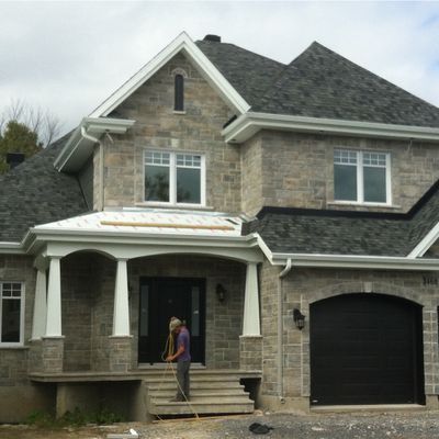 Maison en pierre à deux étages avec garniture blanche et porte de garage noire ; personne balayant les marches d'entrée.