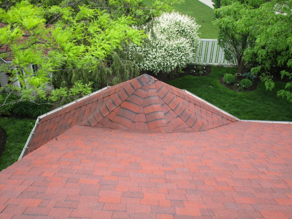 Toit en bardeaux rouges avec pointe pointue, arbres verts et pelouse en arrière-plan.