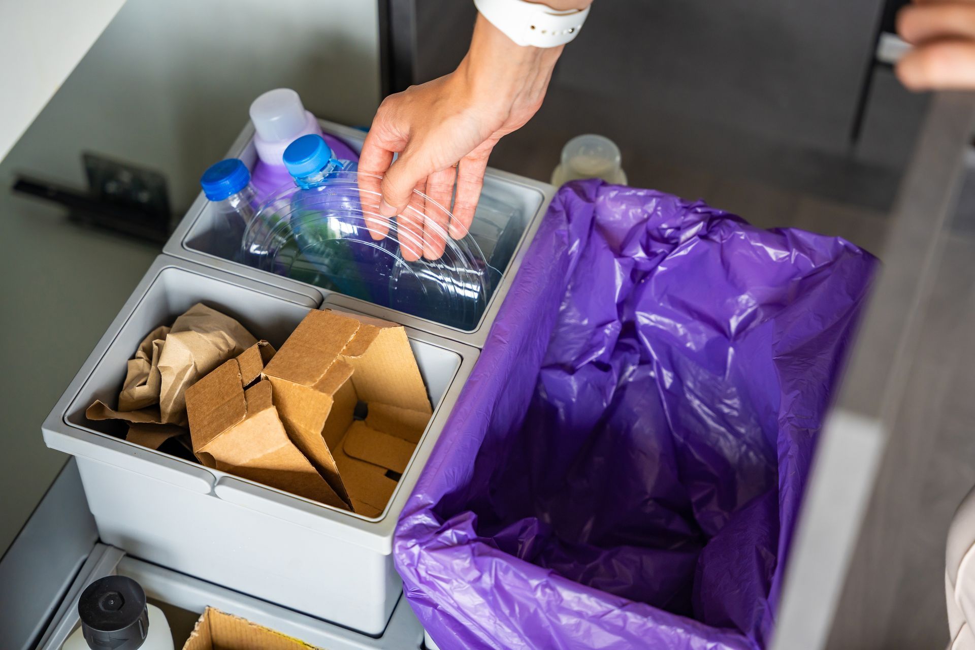 A person is putting a plastic bottle into a purple trash can.