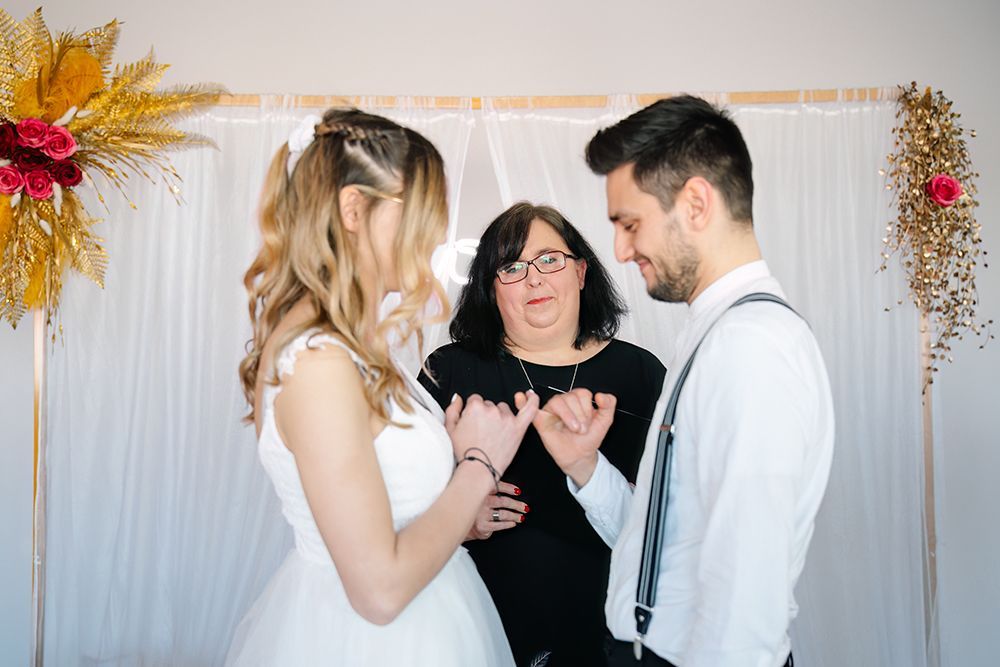 Hochzeitszeremonie im Gewächshaus. Braut und Bräutigam sitzen, dem Trauredner in Hellblau zugewandt. Blumenschmuck.