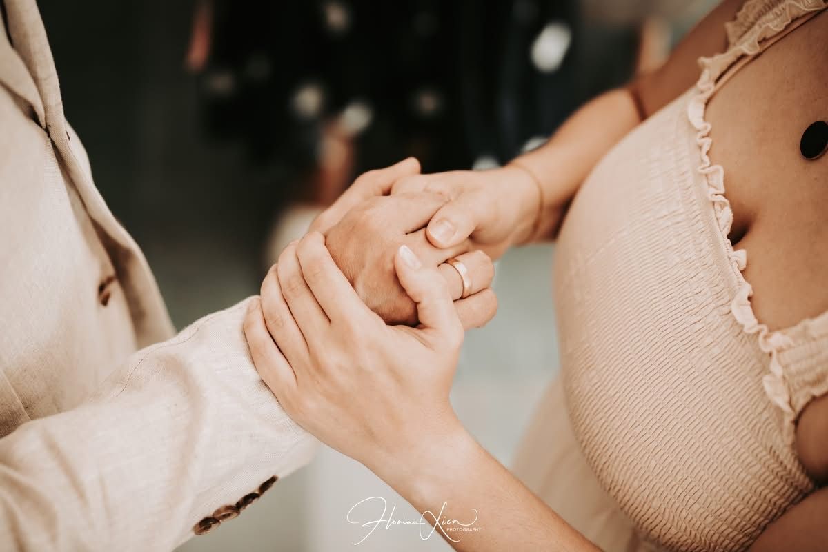 Close-up of hands, one wearing a ring, clasped together. One person in cream-colored top, other in tan suit.