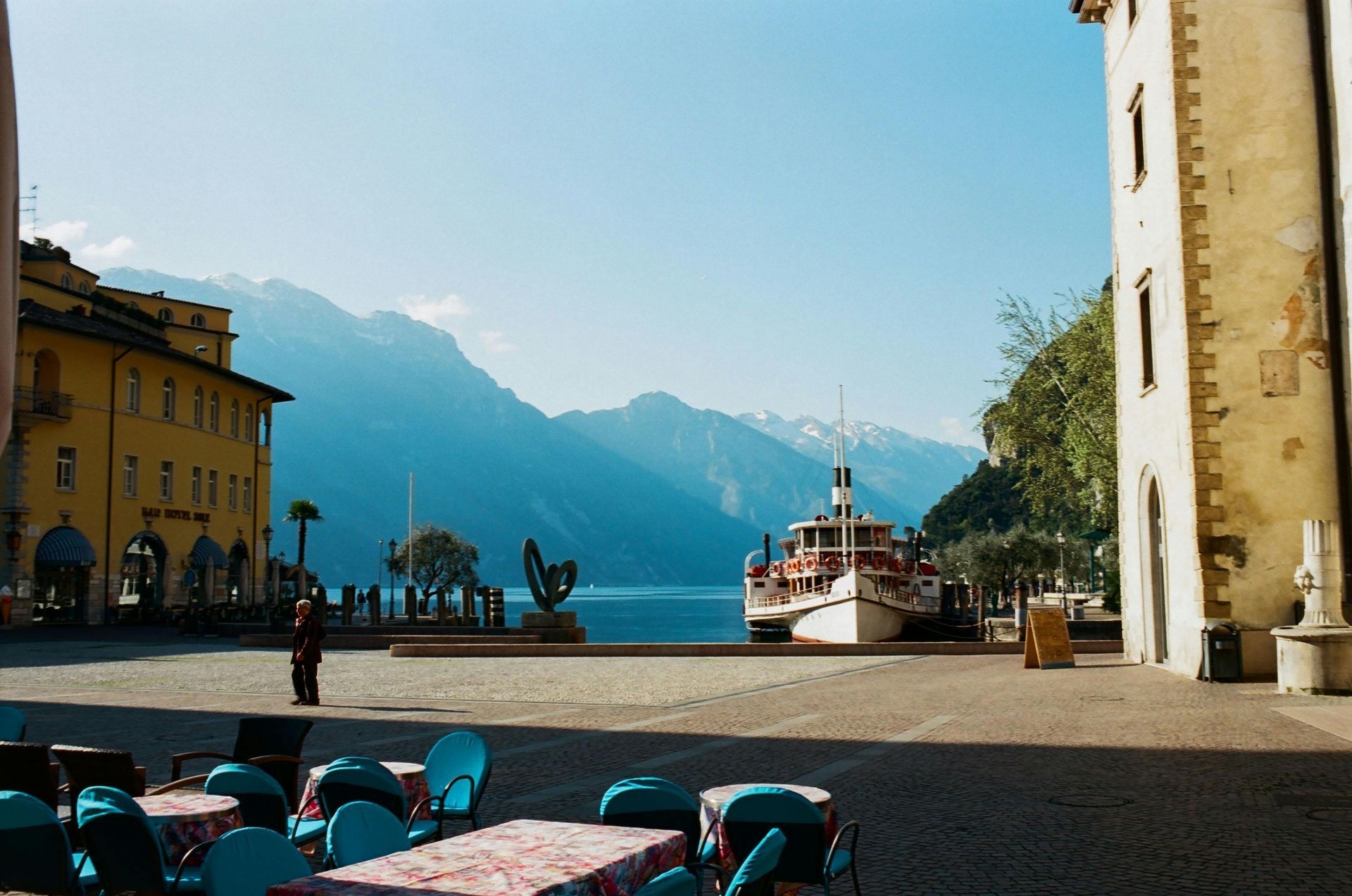 A sunny plaza with a boat docked on a lake, mountains in the distance, and colorful buildings.