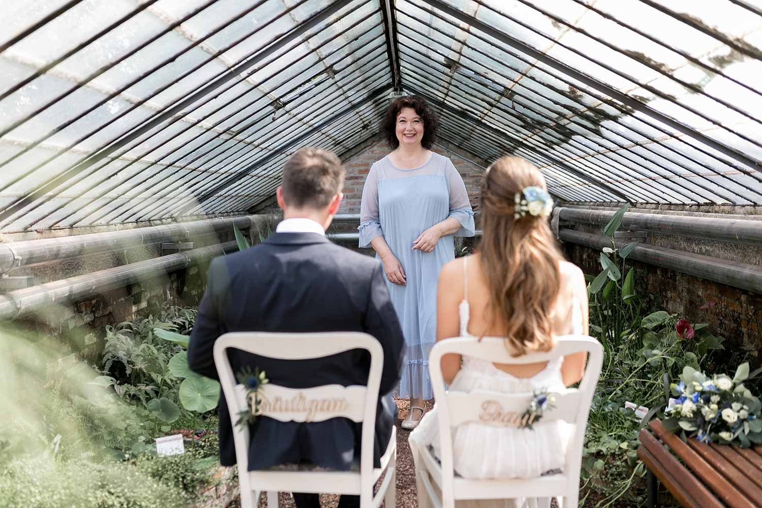 Hochzeitszeremonie im Gewächshaus: Paar sitzt, Traurednerin in blauem Kleid, Blumenschmuck.