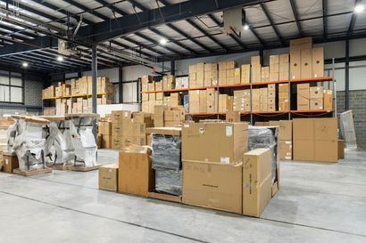 Warehouse interior with stacked cardboard boxes on shelves and the floor, under bright lights.