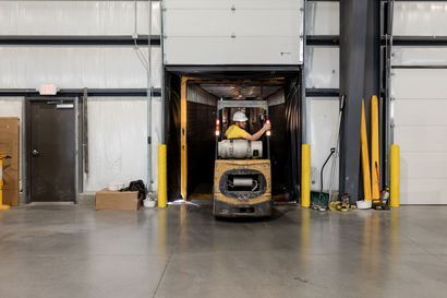 Forklift operator in a warehouse doorway, entering a dock. The operator wears a hardhat and a yellow shirt. The setting is a gray warehouse.