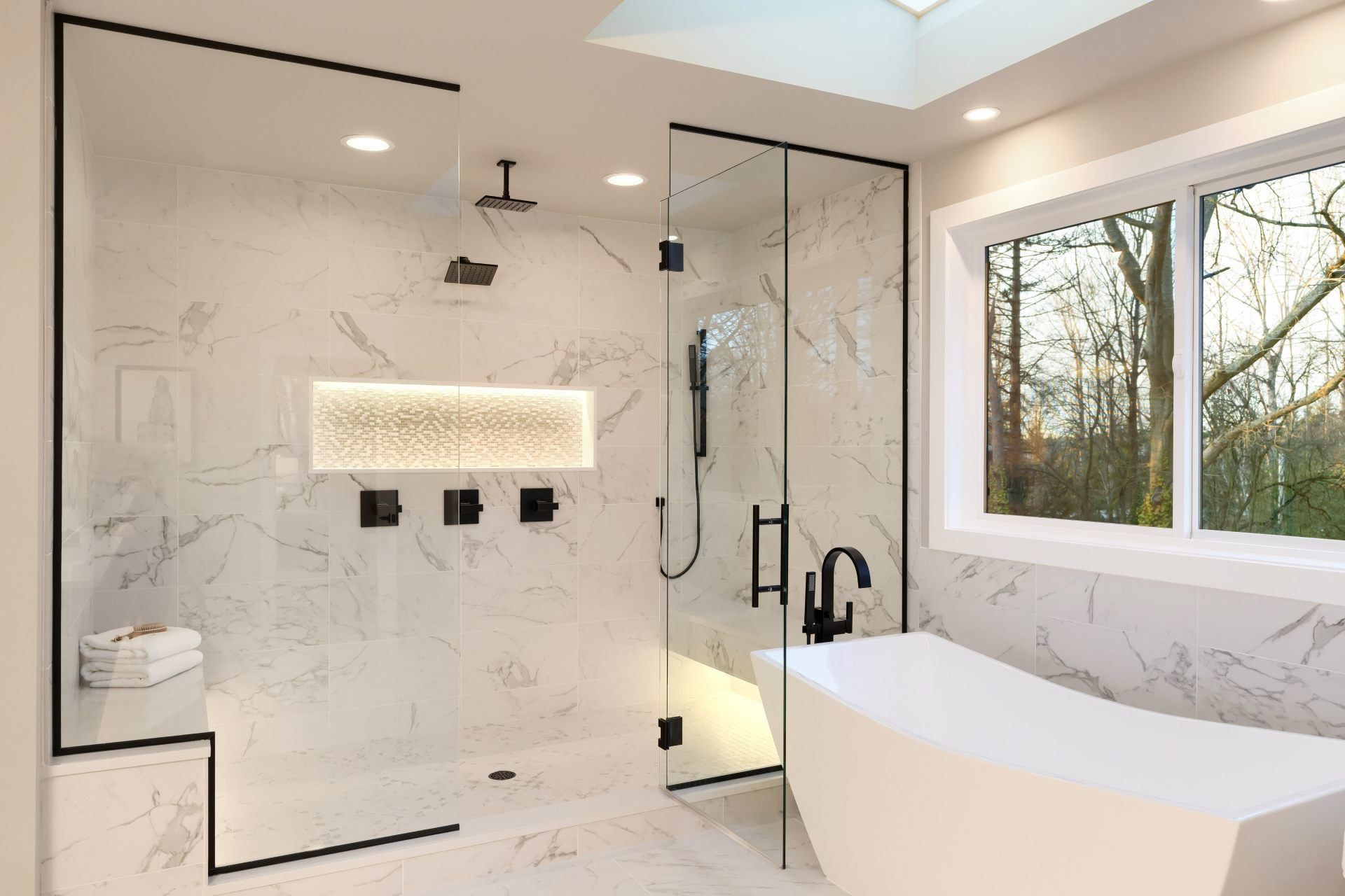 Modern bathroom with a glass-enclosed shower, freestanding tub, marble tile, and a large window.