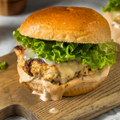 A chicken burger with melted cheese, lettuce, and creamy sauce on a toasted bun, sitting on a wooden cutting board.