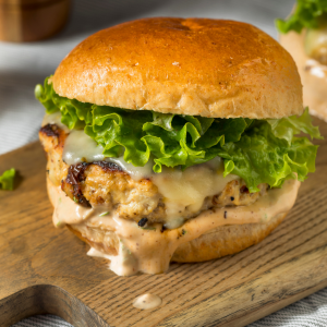A chicken burger with melted cheese, lettuce, and creamy sauce on a toasted bun, sitting on a wooden cutting board.