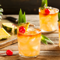 Three rum punch cocktails garnished with pineapple slices and cherries, arranged on a rustic wooden table with fruit.