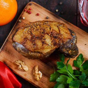 A seared foie gras steak on a wooden board with walnuts, fresh parsley, an orange, and red sauce in the background.